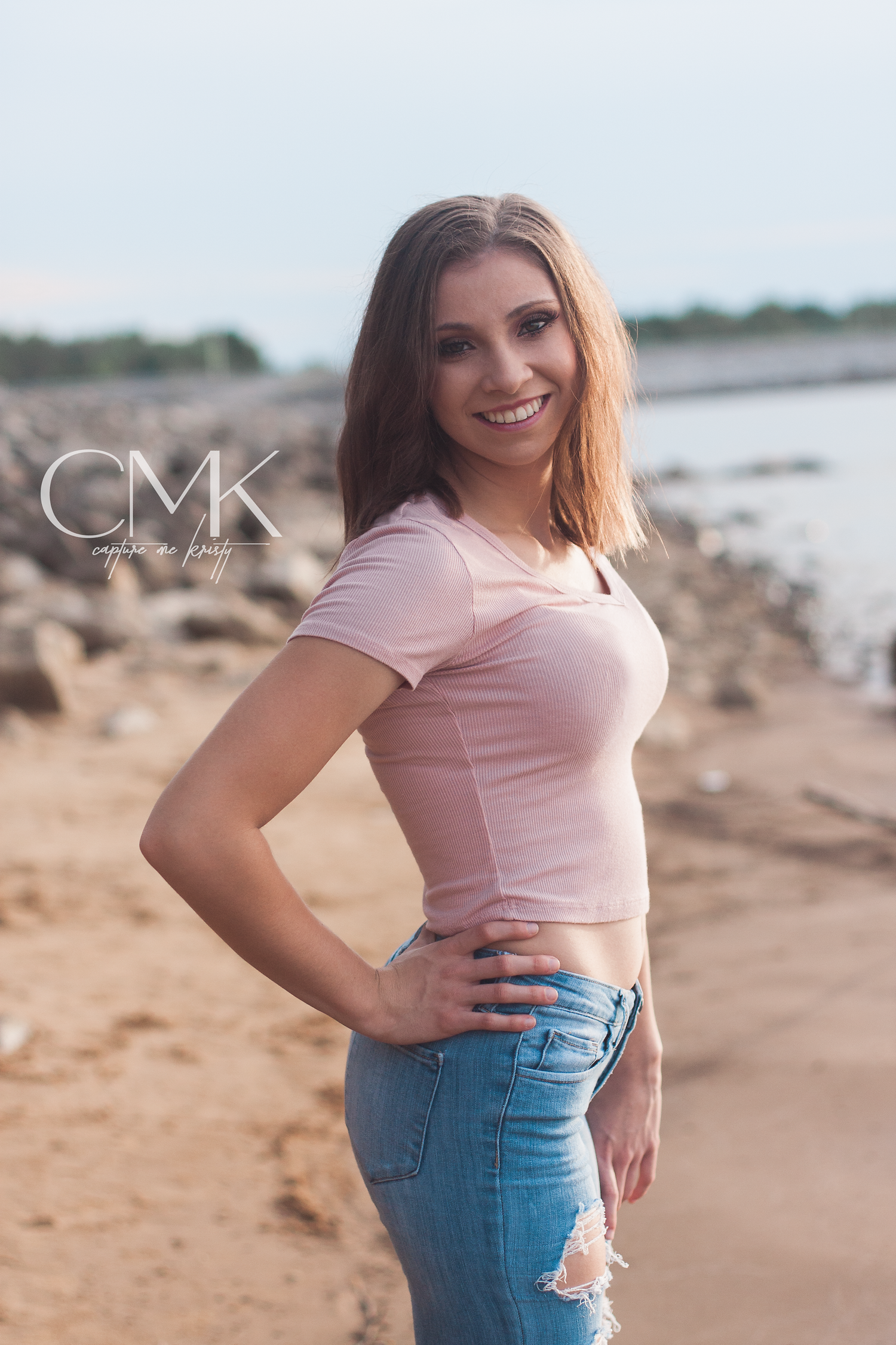 Casual beach portrait in pink crop top and distressed jeans with rocky coastline backdrop.