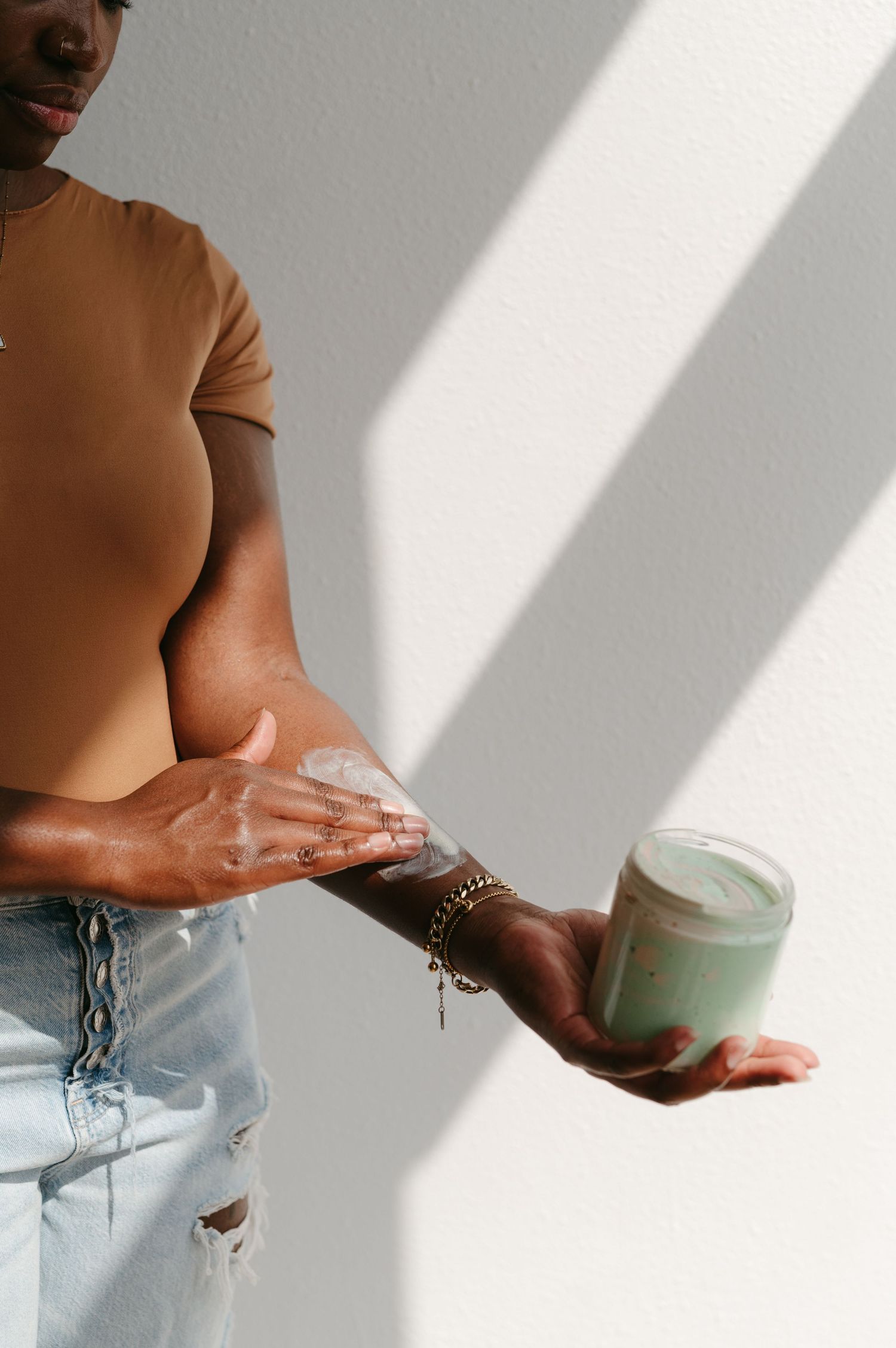 Hands applying moisturizer cream from a mint green jar in natural lighting with dramatic shadows.
