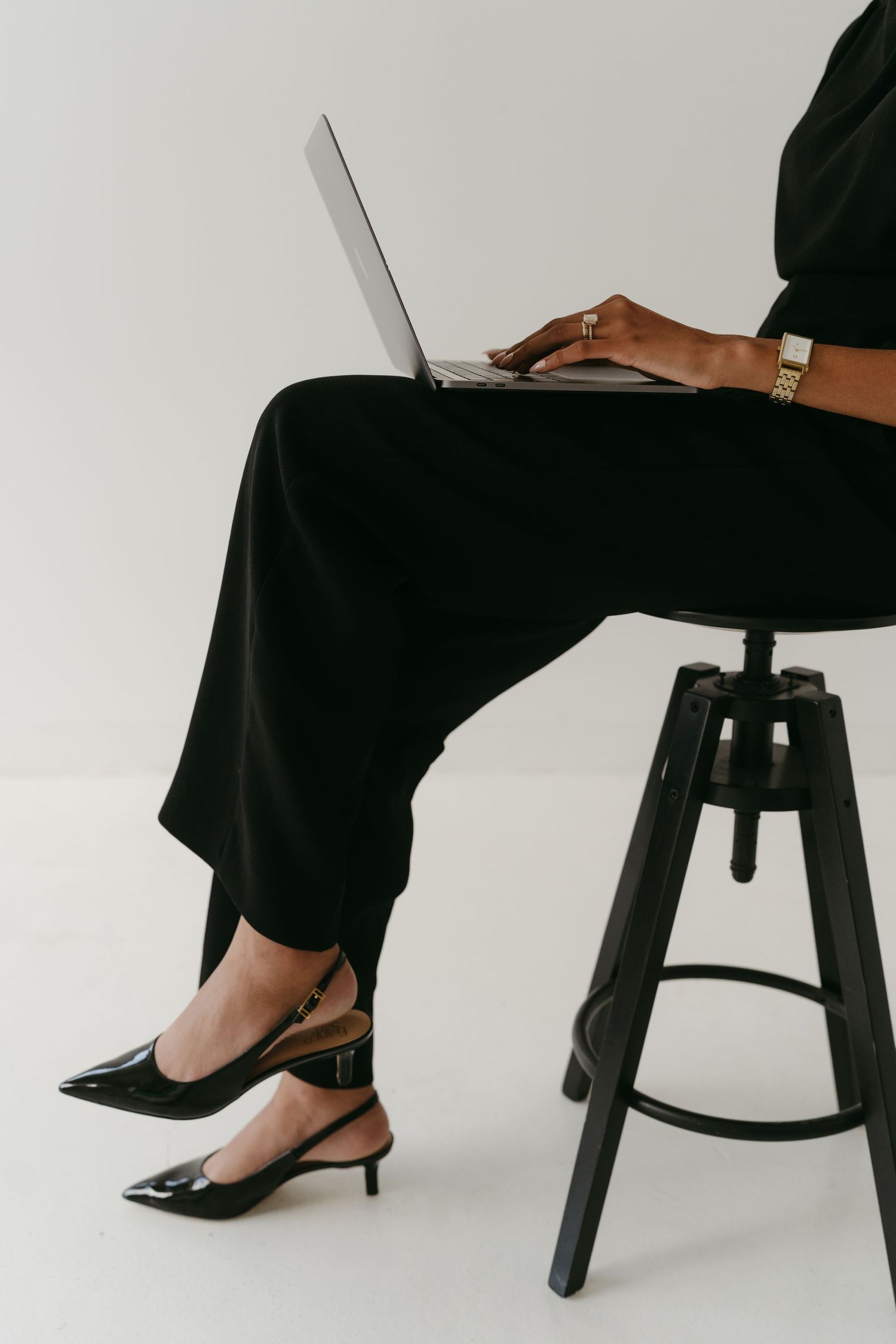 Person working on laptop while sitting on industrial stool, wearing elegant black attire.
