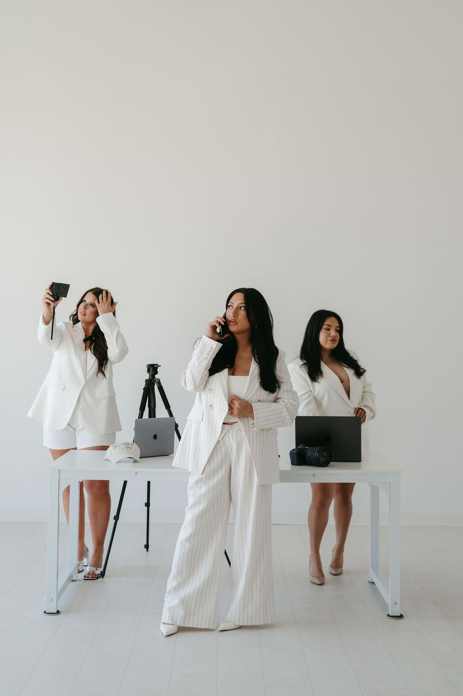 Three women in white suits pose with laptops and phones at a modern white desk in a bright studio.