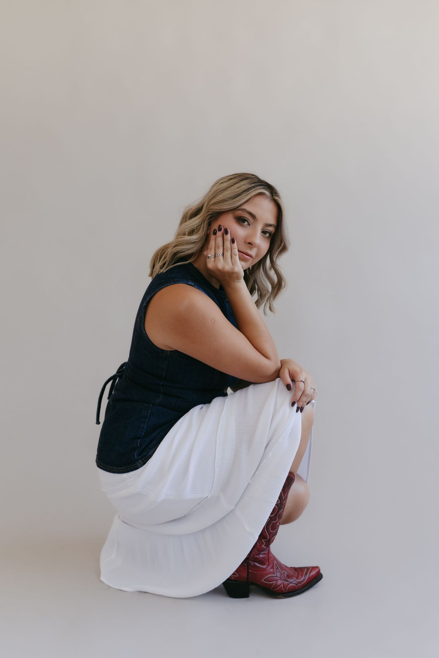 Model in black top and white dress sits contemplatively on floor in minimalist setting.