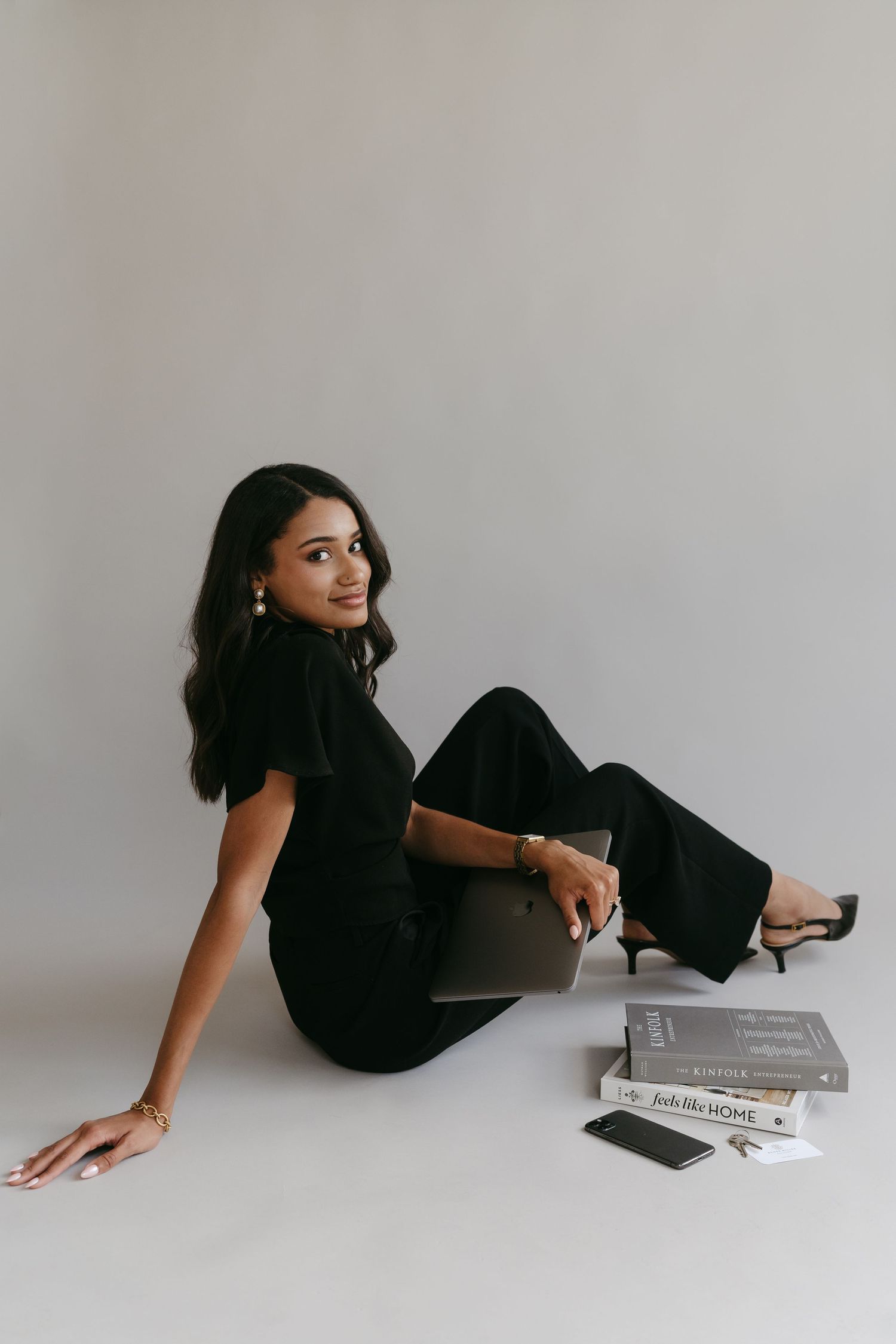 Person in black outfit reclining on floor with books and magazines in minimalist setting with natural light.