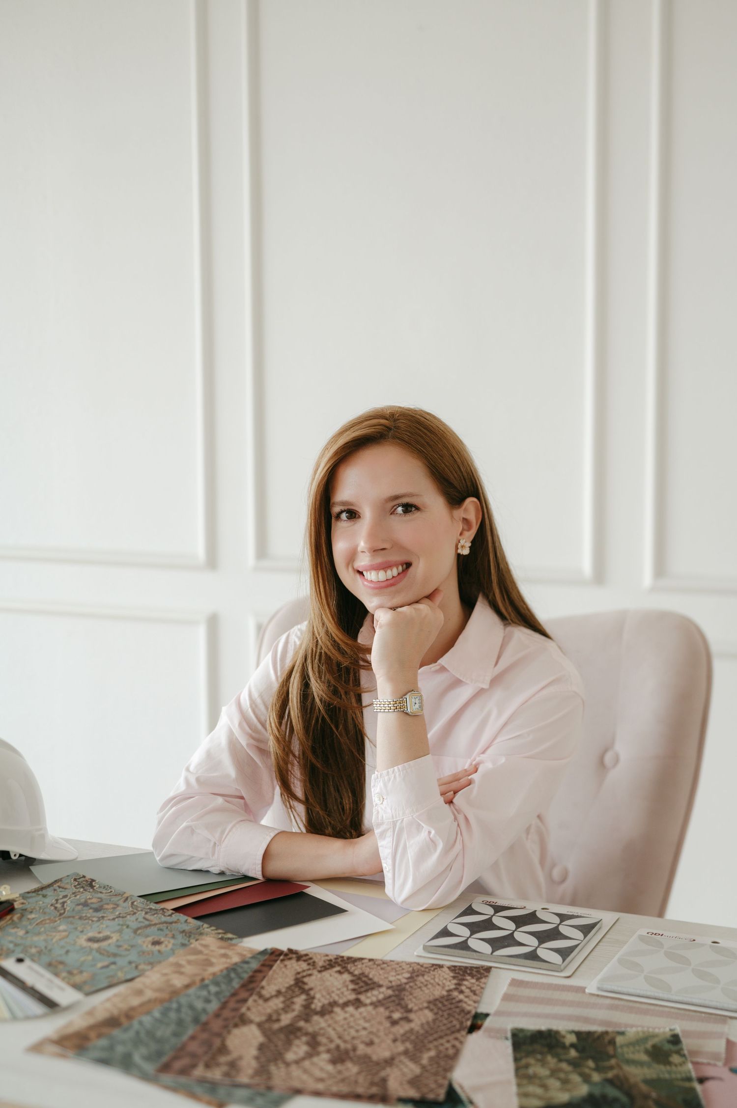 Smiling woman in pink shirt sits at desk with fabric swatches and design samples in a bright white office.