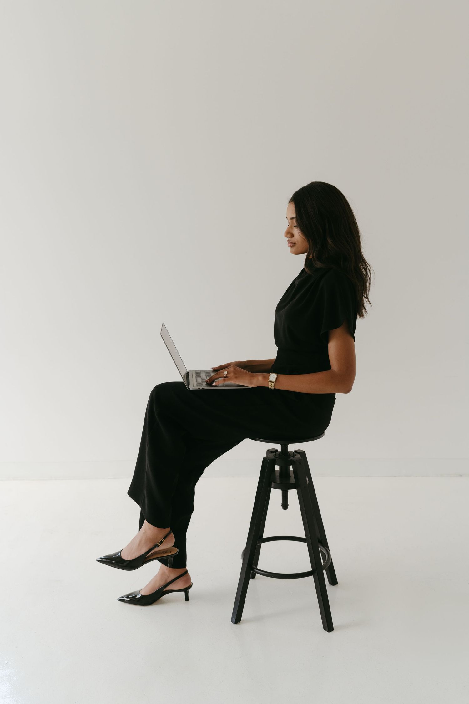 Professional woman in black outfit working on laptop while sitting on modern chair against white background.