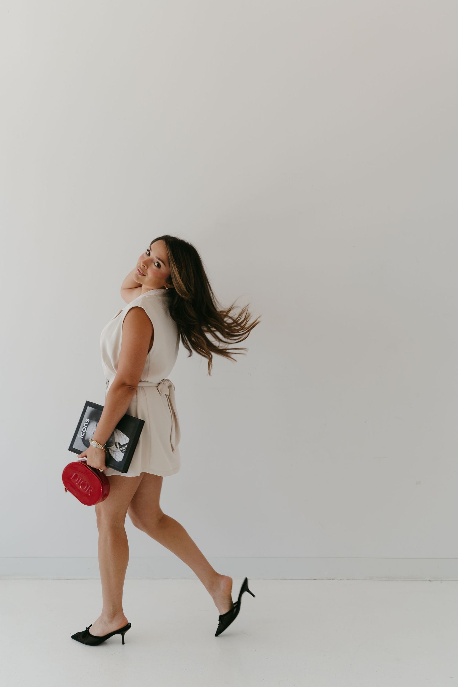 A figure in a white dress twirls playfully while holding accessories against a minimalist white background.