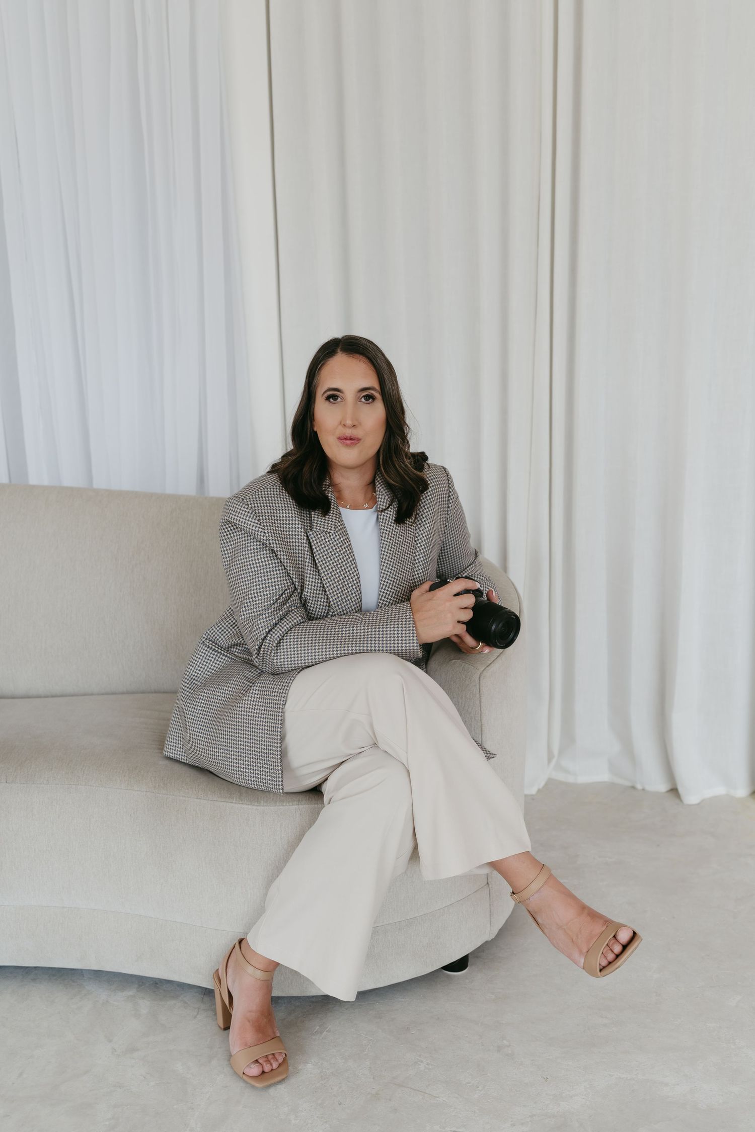 Model in grey blazer and white pants reclines in minimalist studio setting.