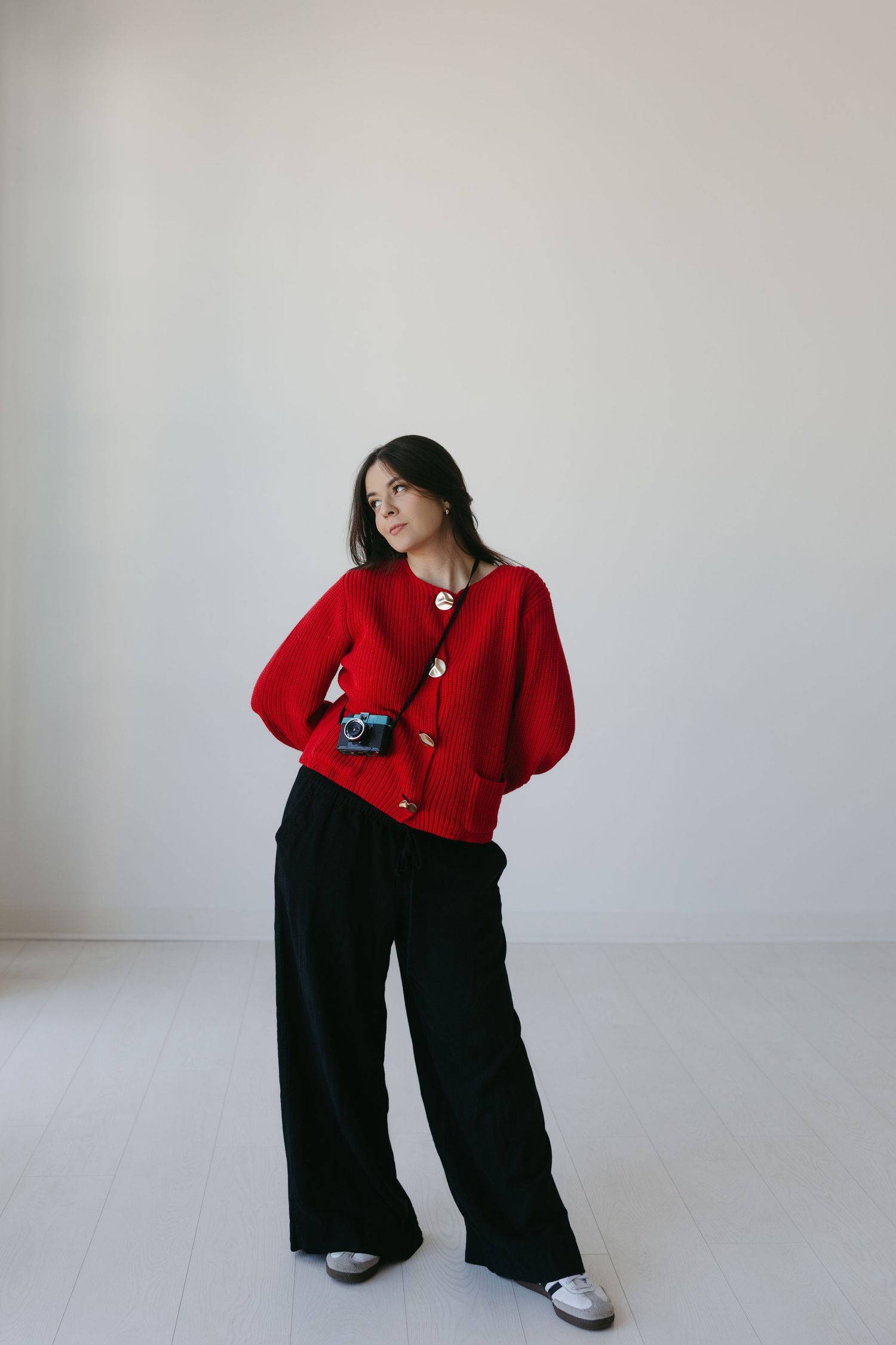 Woman in red cardigan and black wide-leg pants poses with a vintage camera in a minimalist white room.