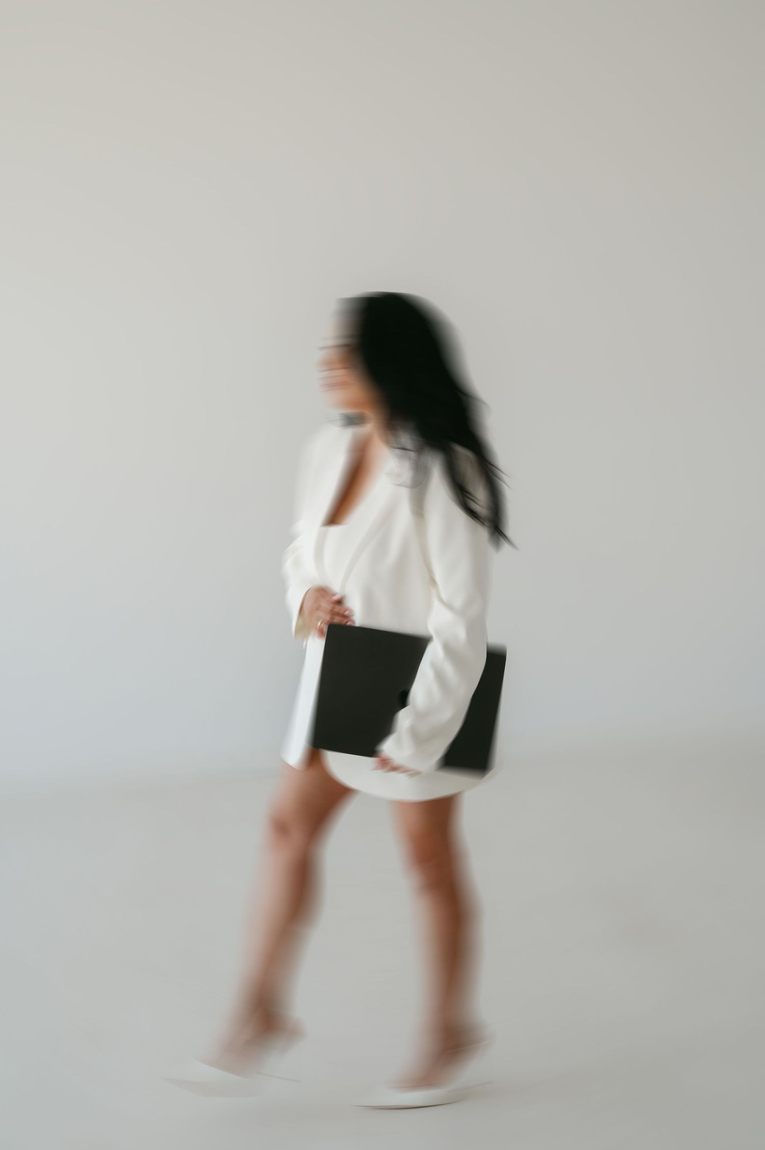 A woman in a white blazer and skirt walks with a black laptop in a minimalist white studio setting.