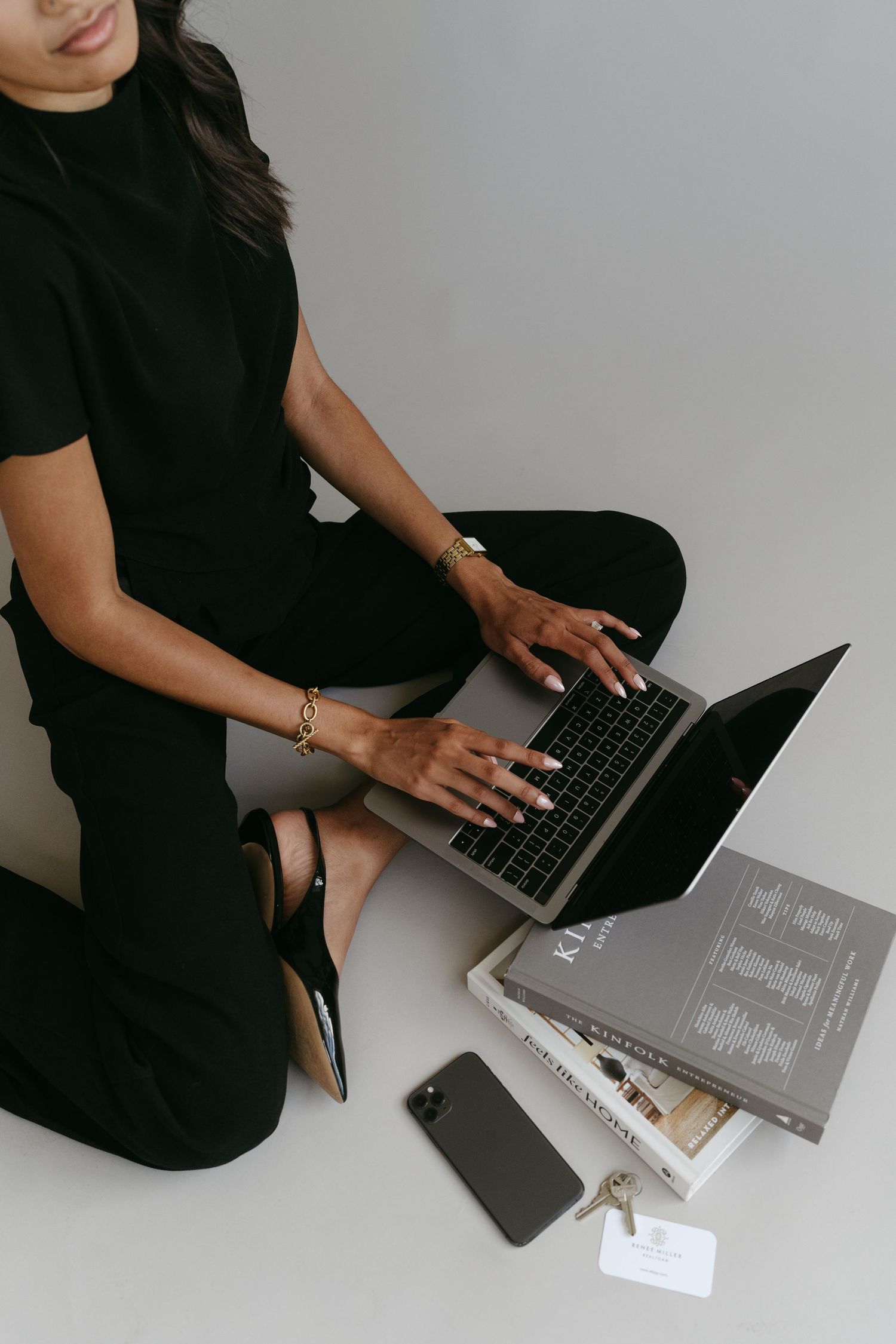 Overhead view of woman typing on laptop with books and phone nearby on white surface.