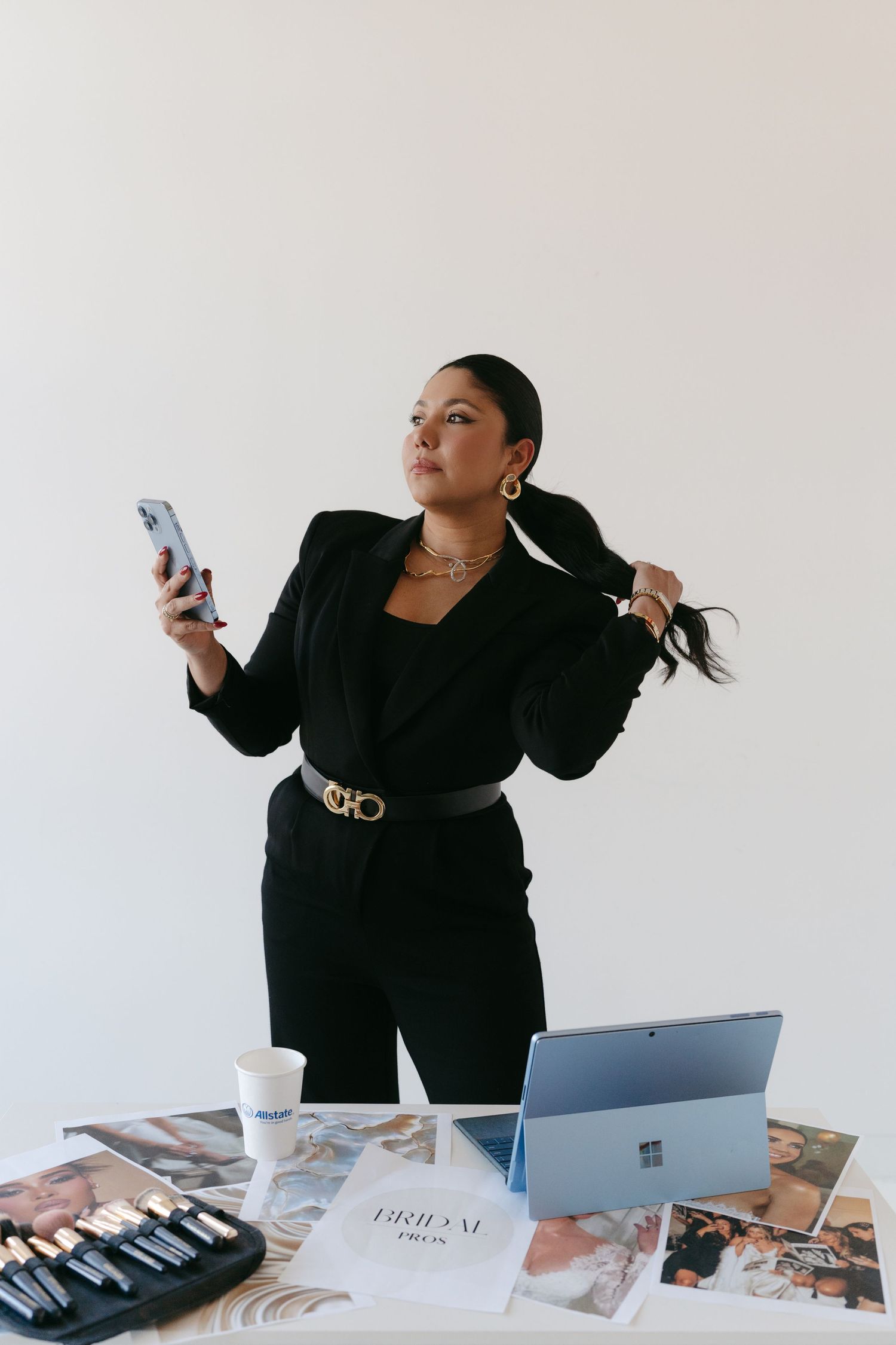 Professional in black business attire checking phone while standing at desk with laptop and marketing materials.