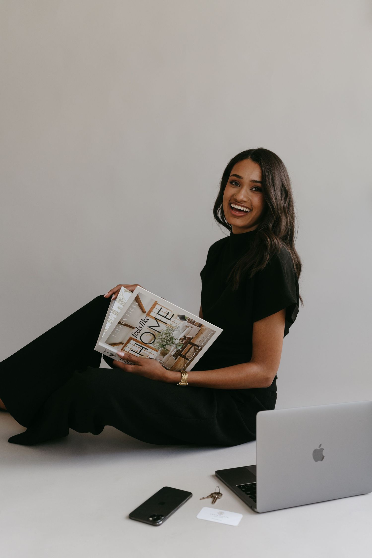 Professional woman in black outfit smiling while holding magazine and sitting by laptop.