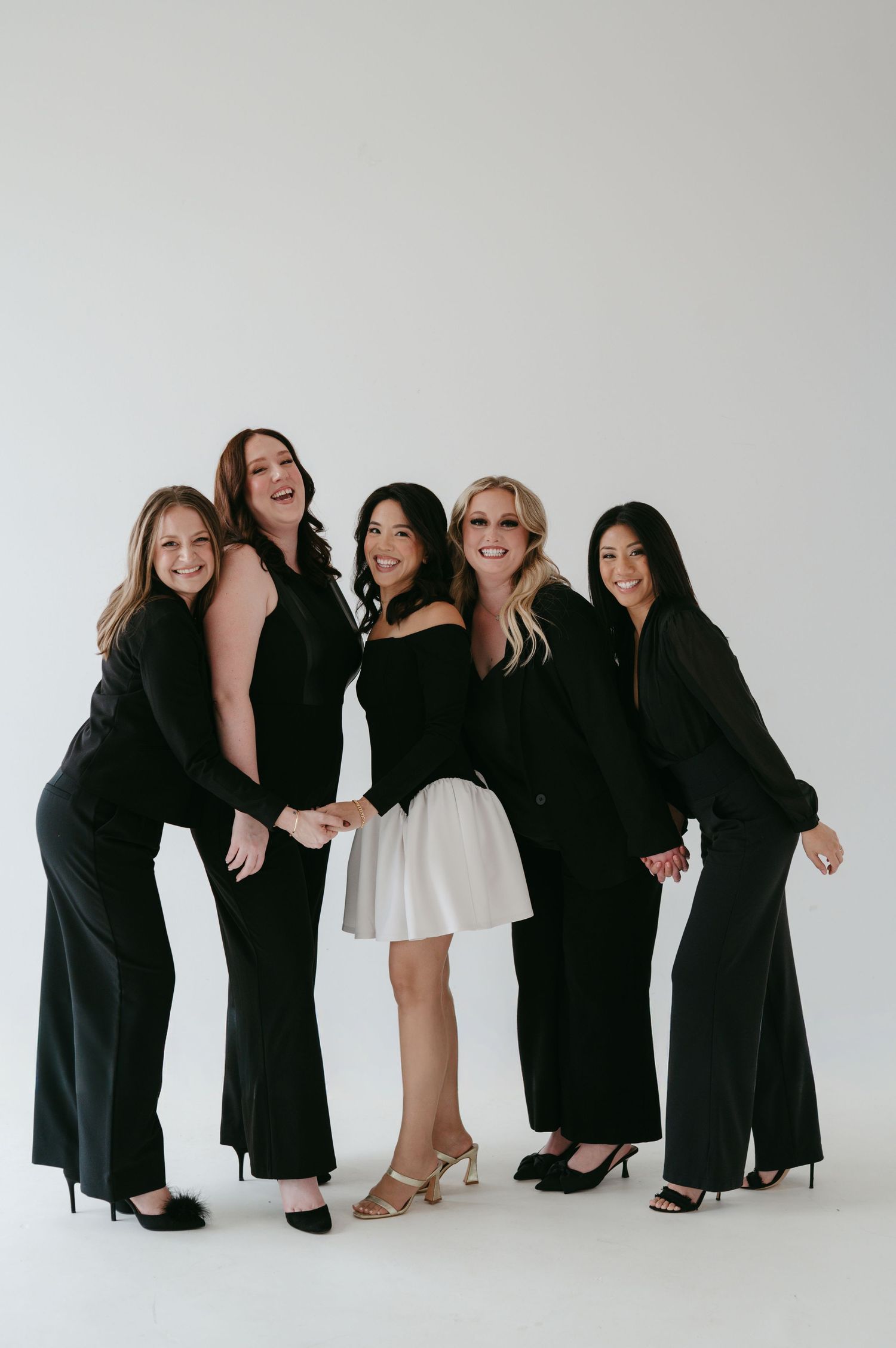 A group of five women posing together in black formal dresses and one white dress against a white studio backdrop.