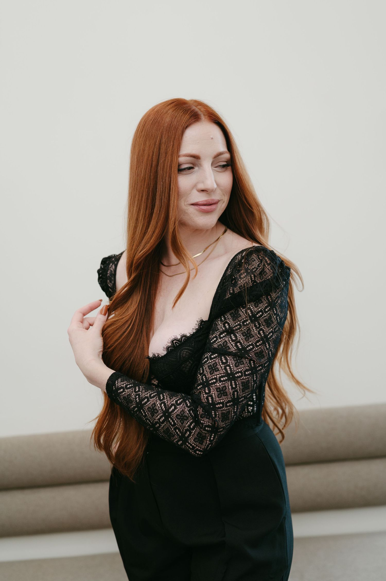 Woman with long red hair wearing a black lace sleeve dress poses against light background.