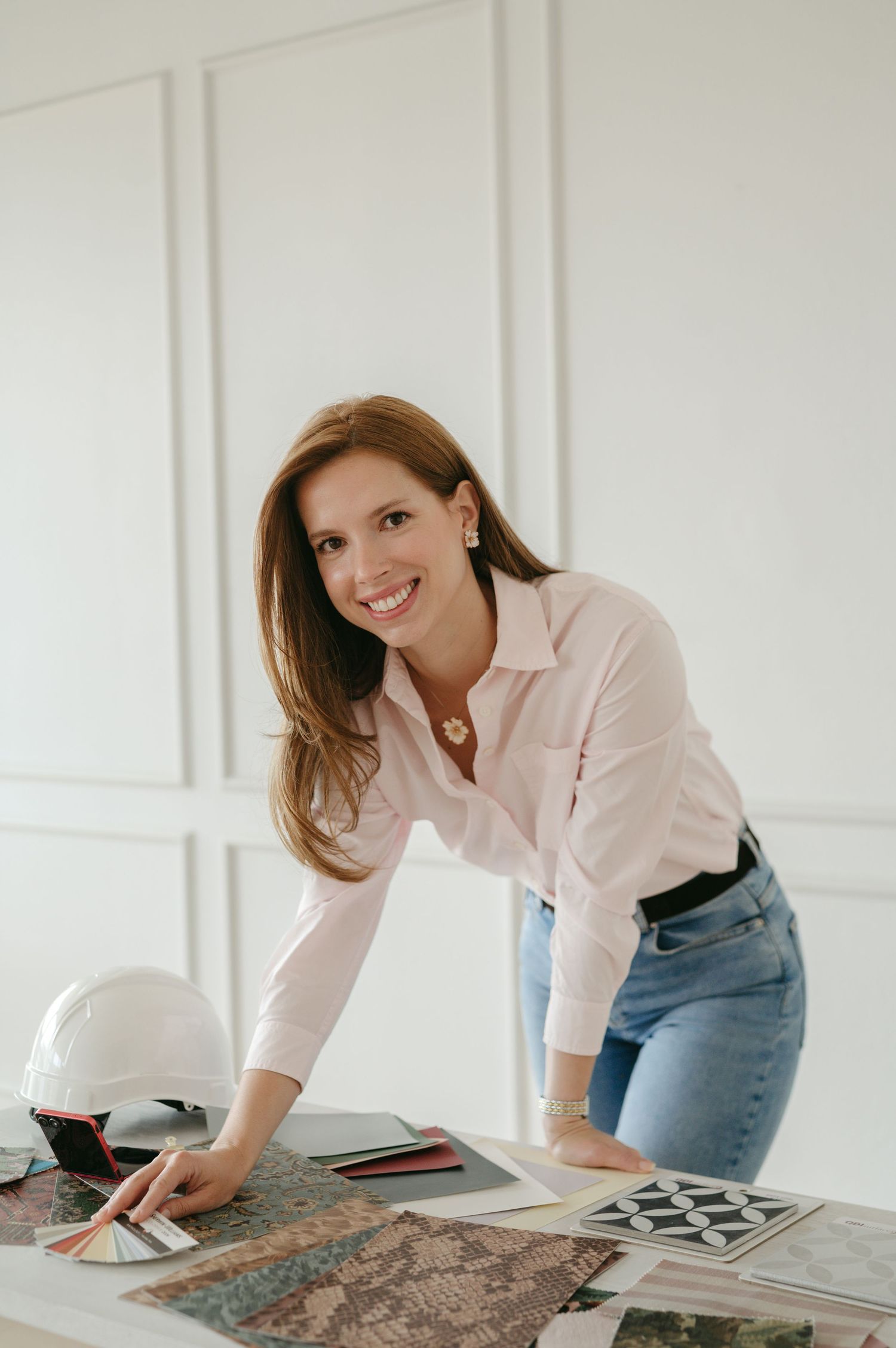 Smiling woman in white shirt and jeans leans over desk with color swatches, blueprints, and hard hat in bright studio.