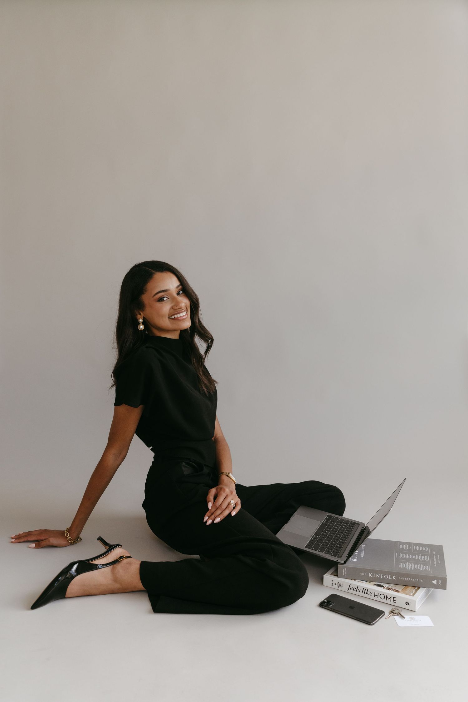 Stylish woman in black outfit sitting casually with laptop and magazines on white floor.