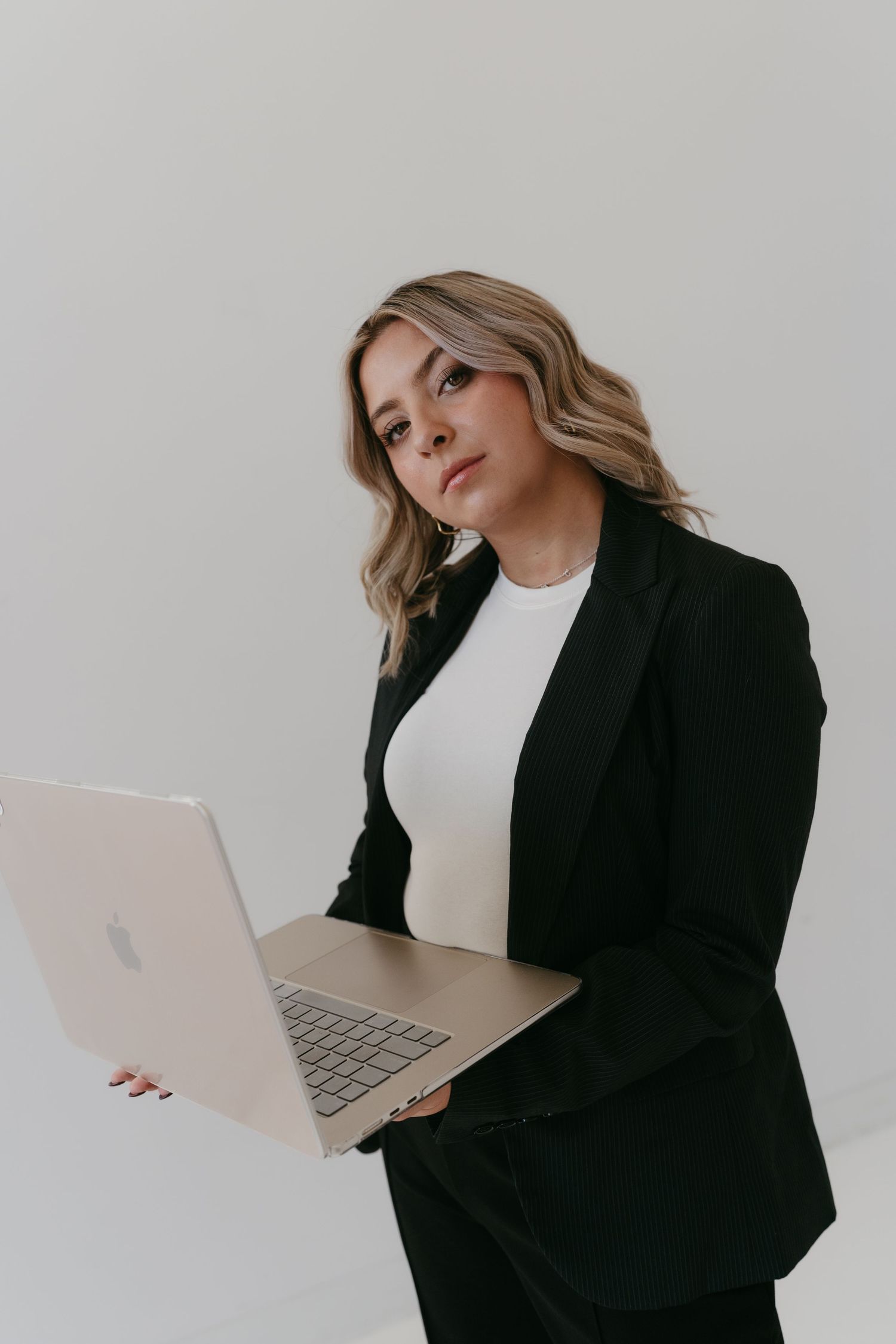 Professional poses with laptop in black blazer against minimal white background.