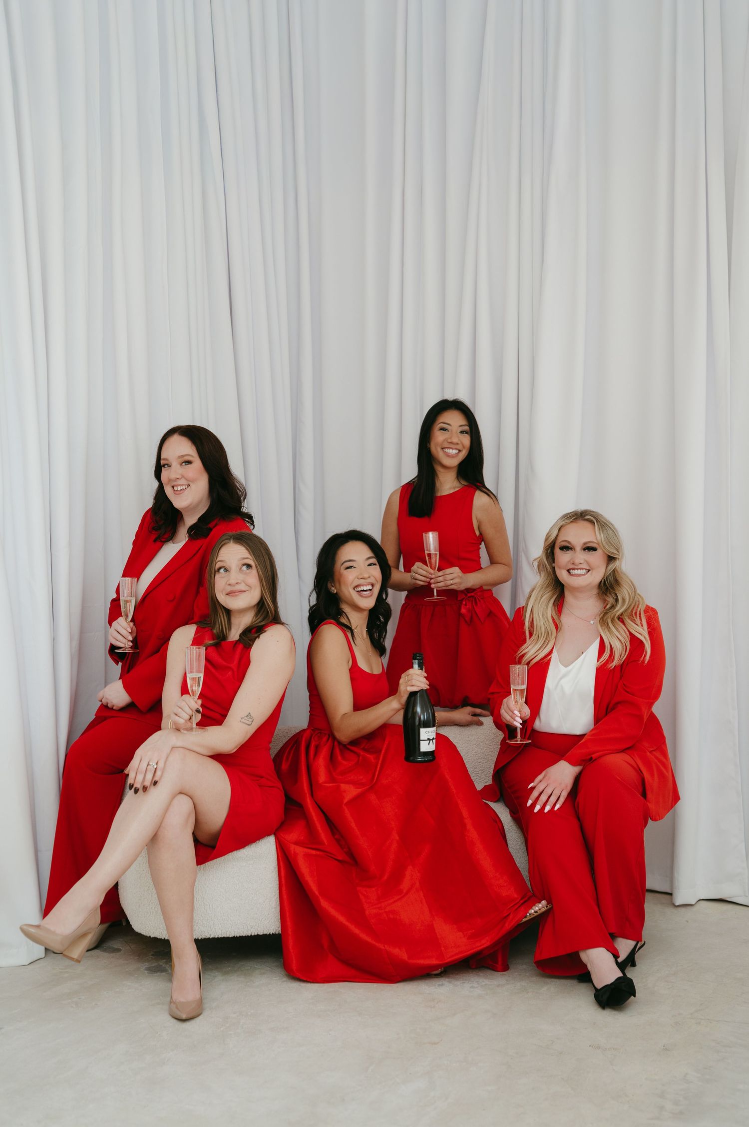 A group wearing red dresses and outfits sits casually against a white curtained wall.