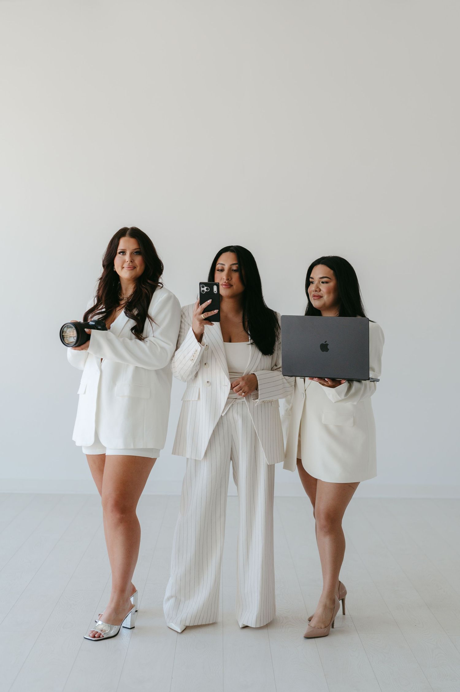 Three women in white blazer outfits pose holding a camera, smartphone, and Apple laptop against a white background.