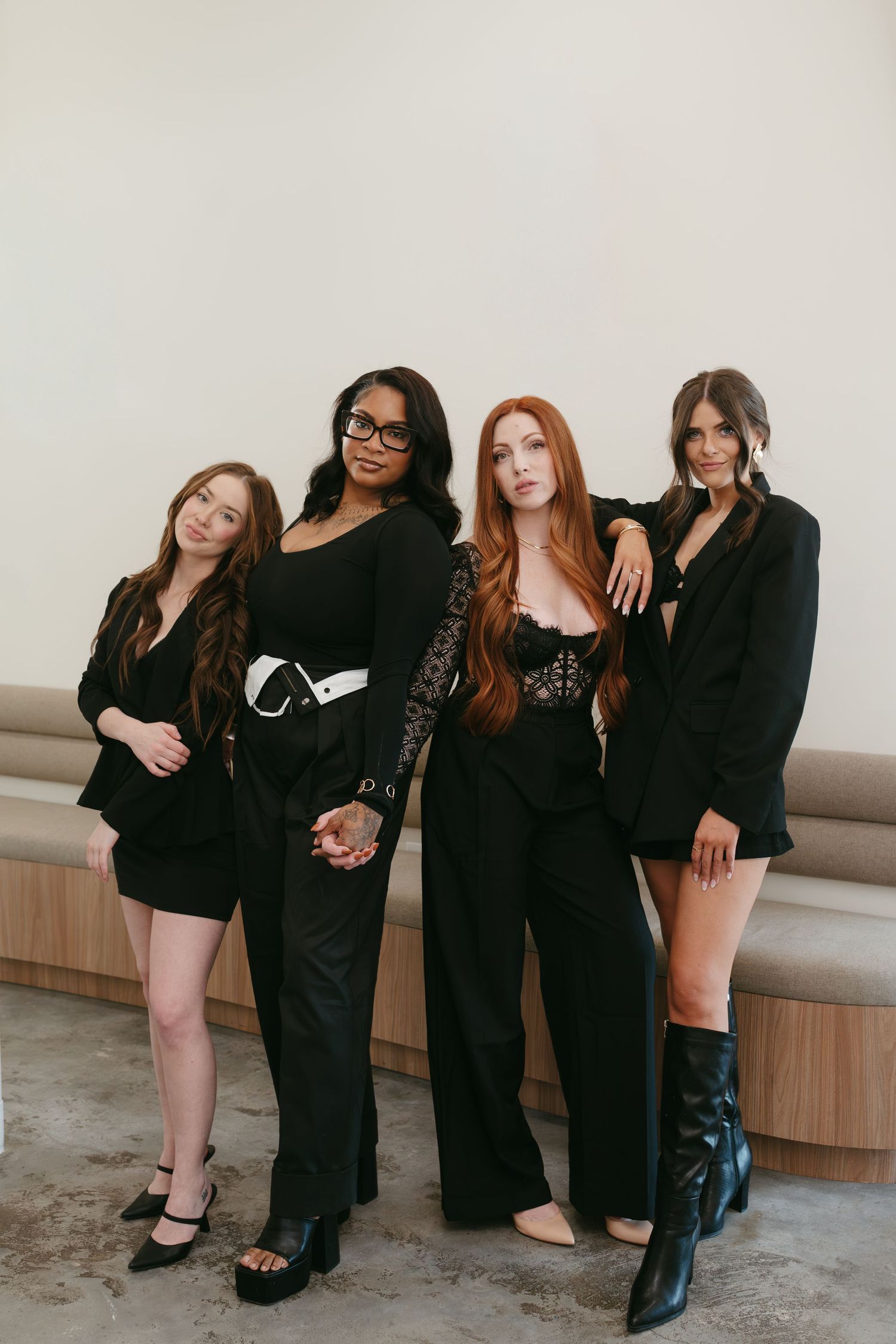 Four women in black outfits pose together for a fashion photo series showing different angles and poses.