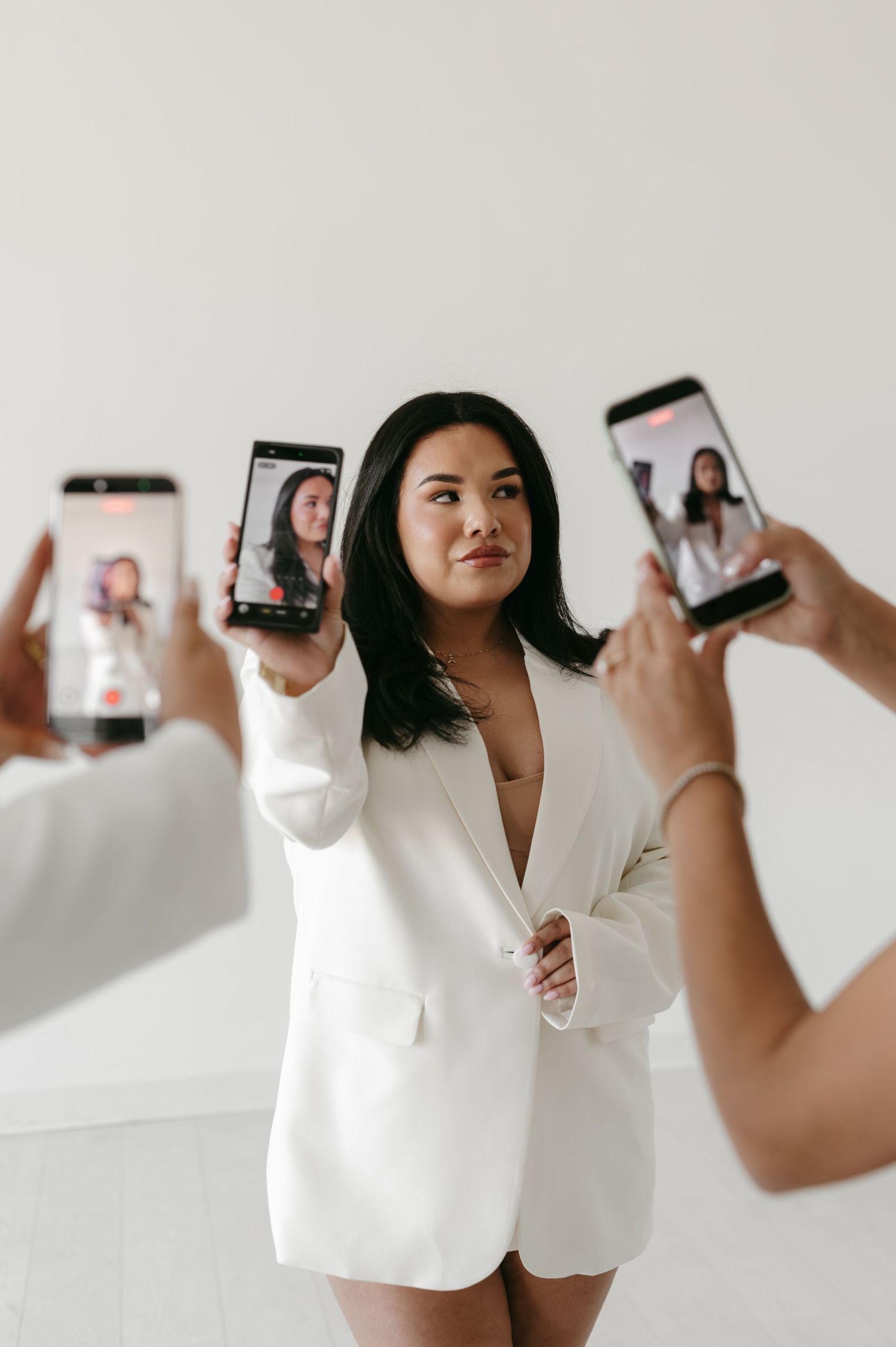 Woman in white blazer holds smartphone while two others photograph her against a white background.