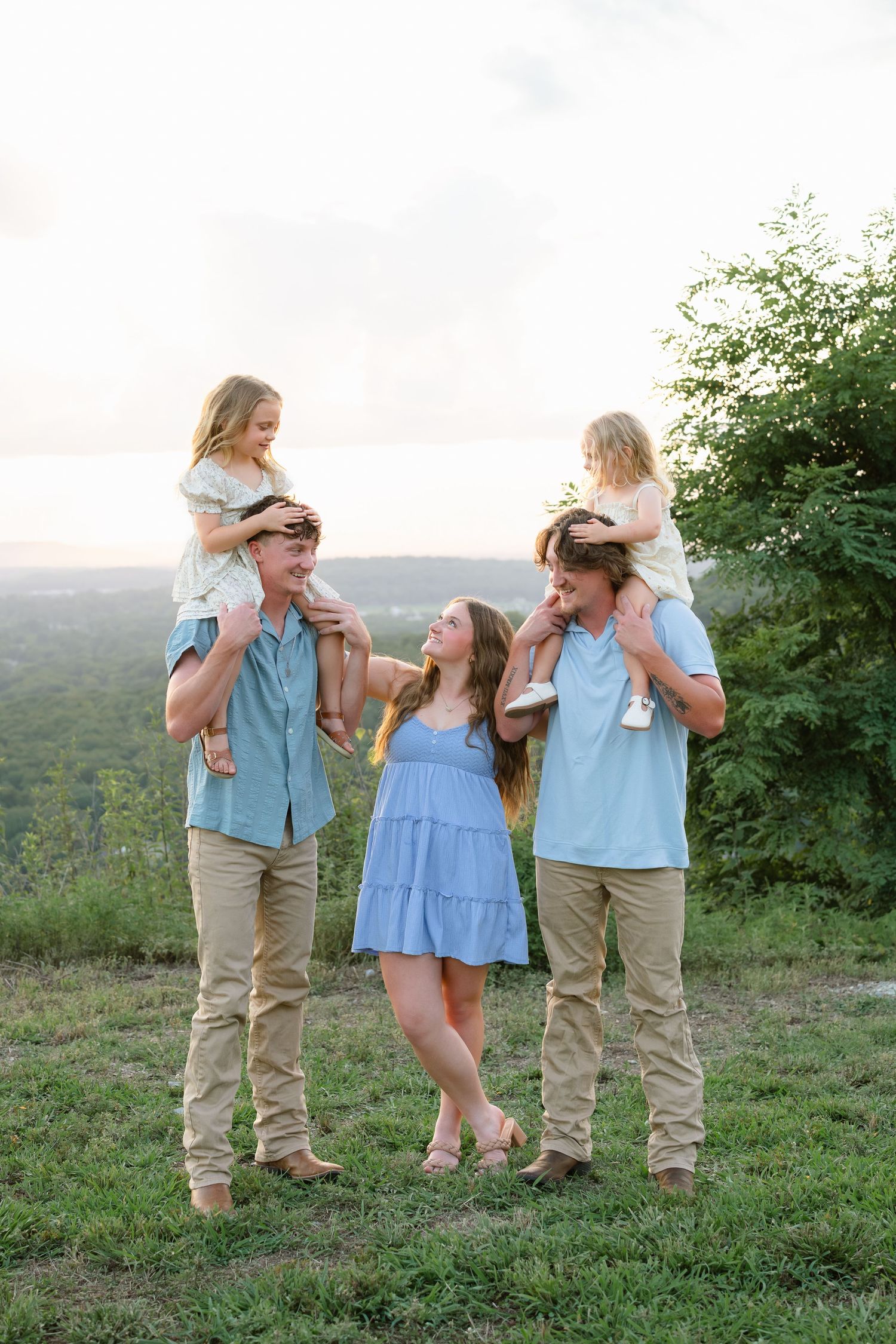 Family of five posing outdoors at golden hour, two children on shoulders of adults in coordinating blue and khaki outfits.