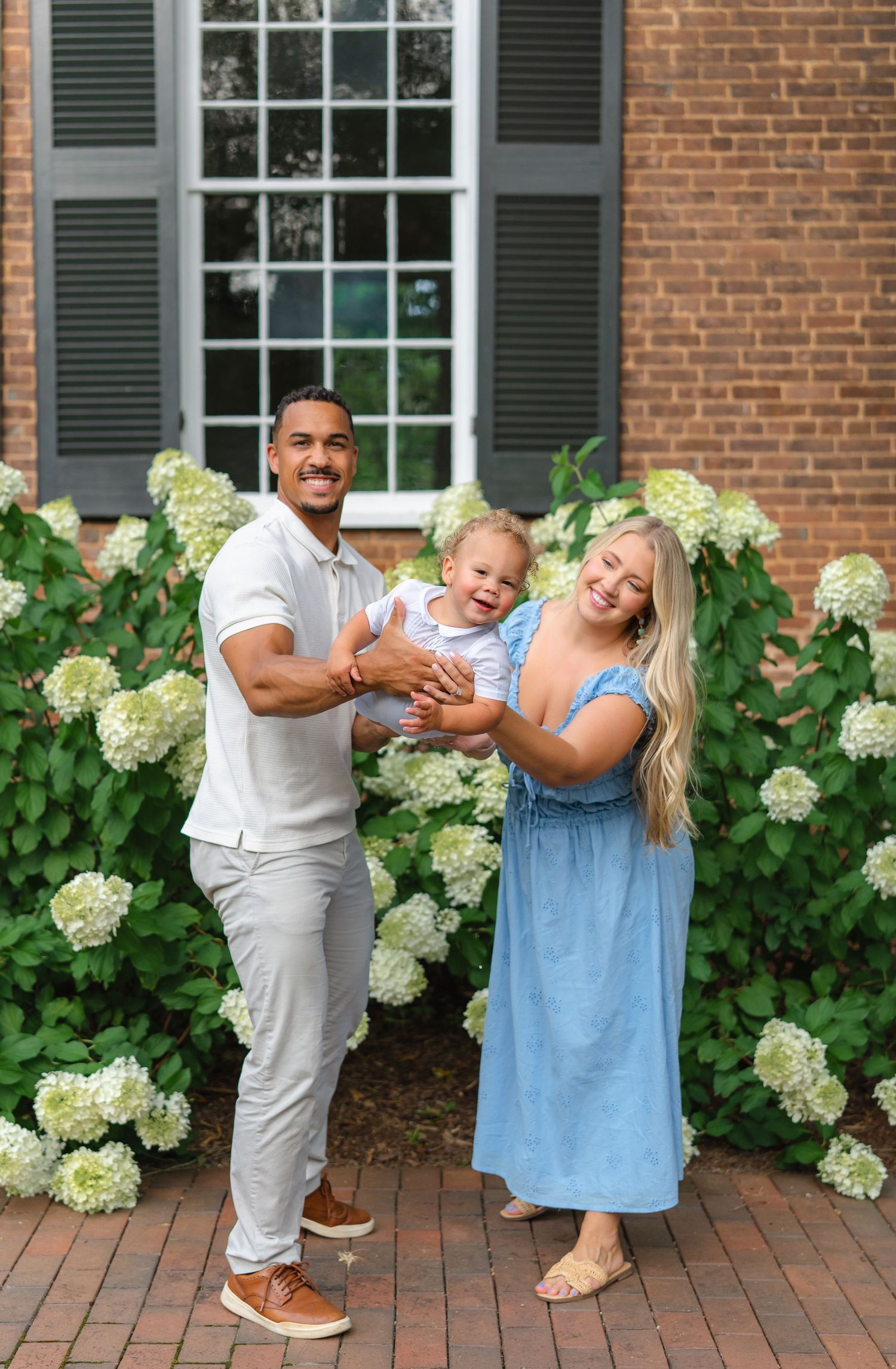 Happy family with a baby posing in front of blooming white hydrangeas and a brick building.