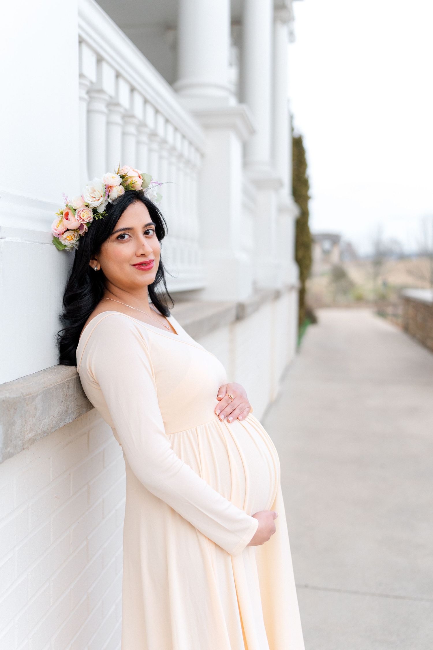Smiling pregnant woman in cream dress and floral crown poses by white columns.