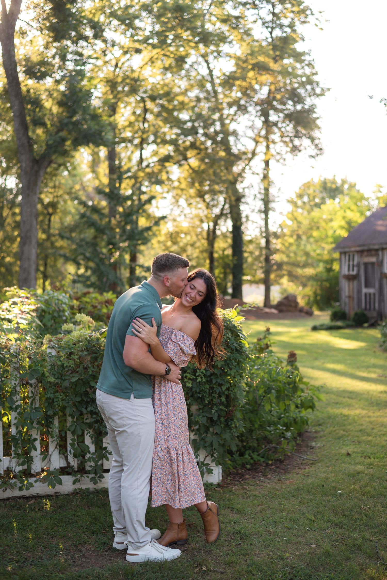 Man kissing woman as she laughs joyfully beside a white fence in a sunny garden.