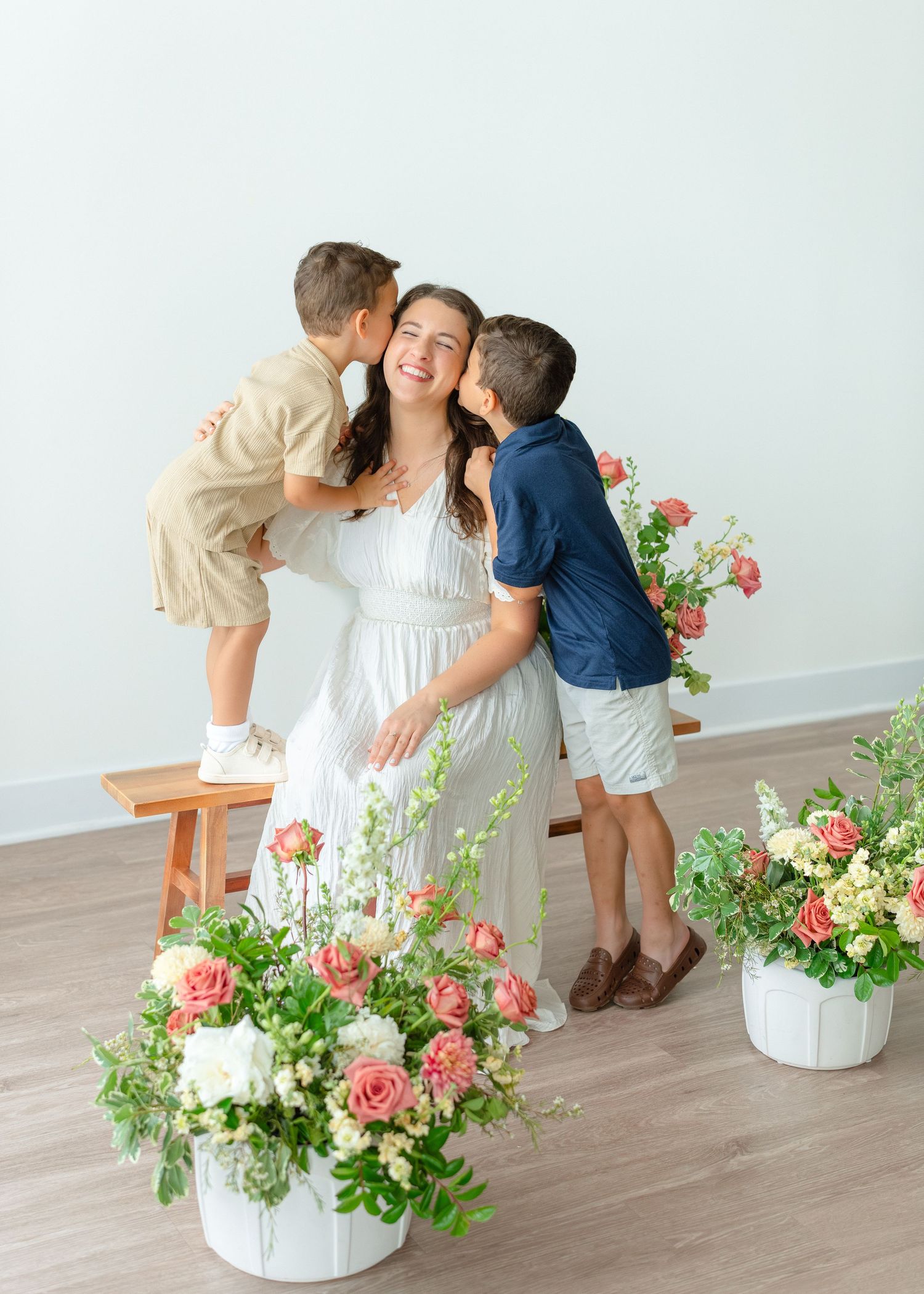 A joyful mother in a white dress laughs as her two young sons kiss her cheeks in a flower studio.
