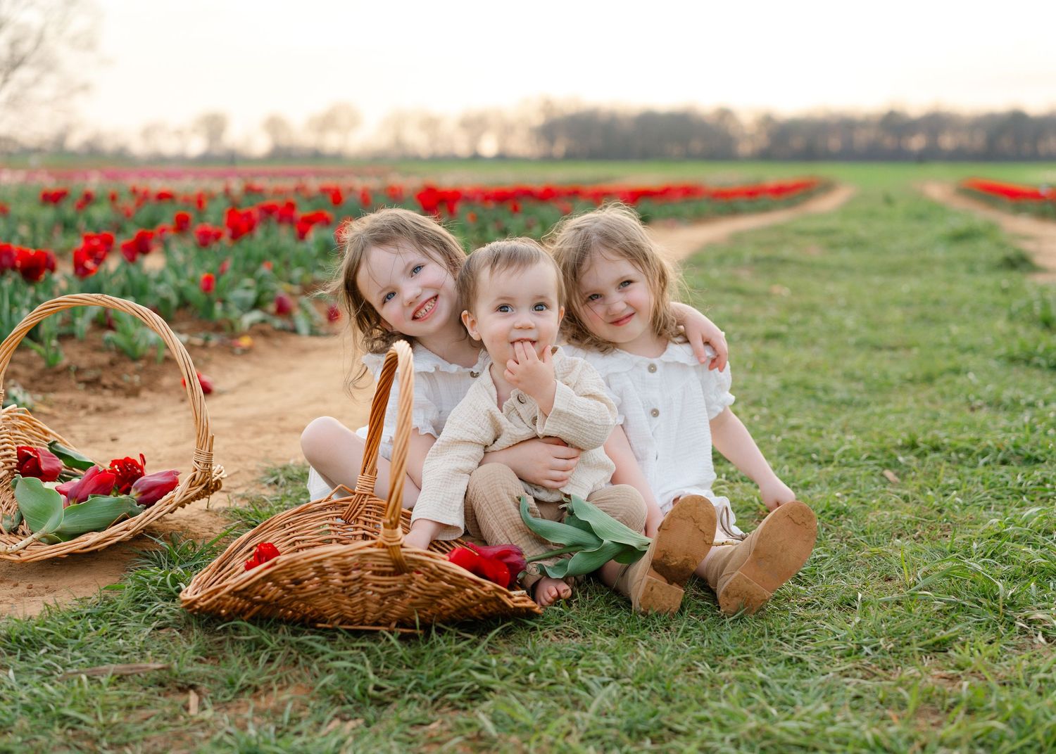 Three young siblings sit smiling in a red tulip field with a wicker basket of flowers nearby.