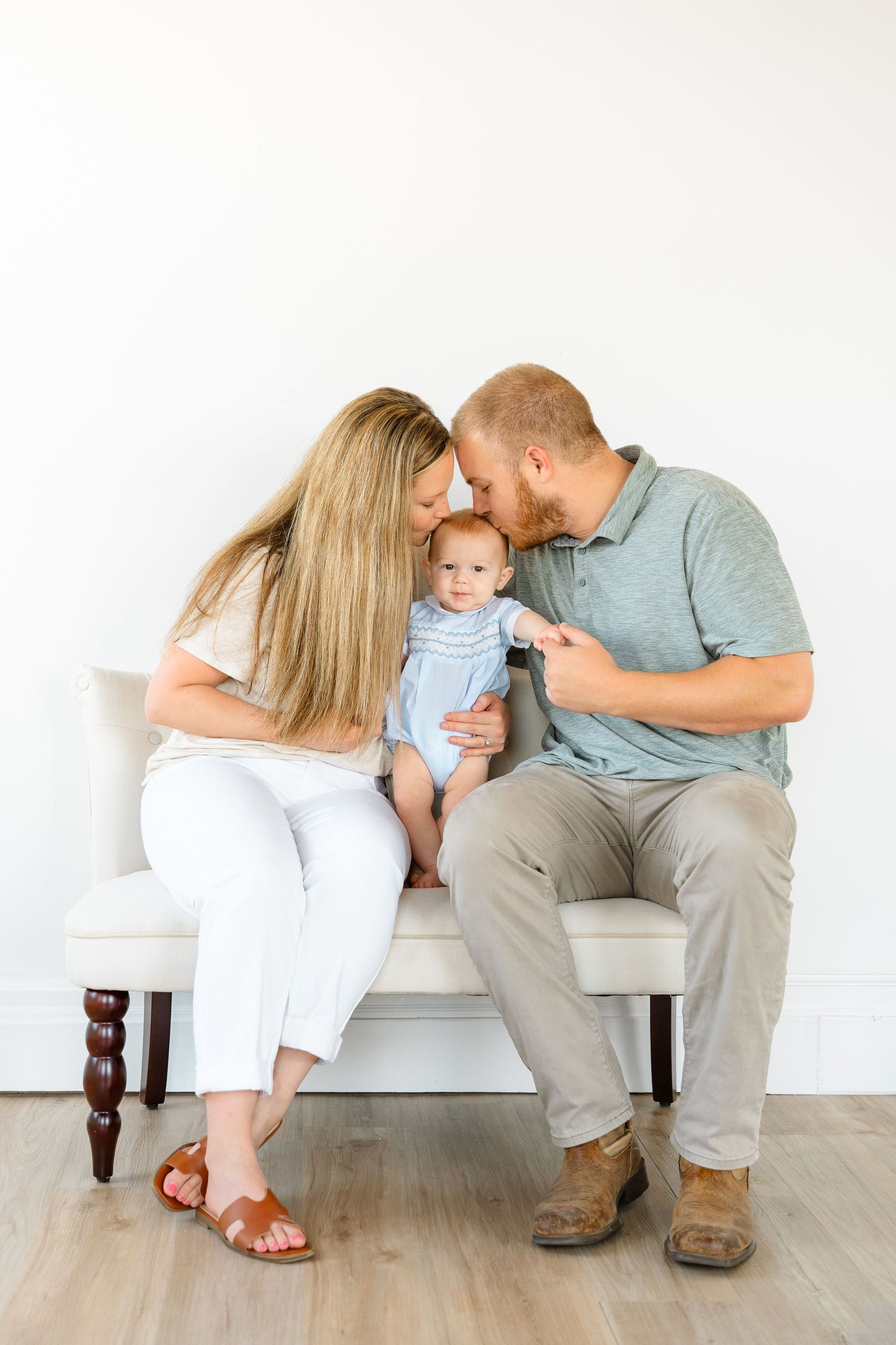 A young couple sits on a white bench, leaning in to kiss their newborn baby dressed in a blue outfit.