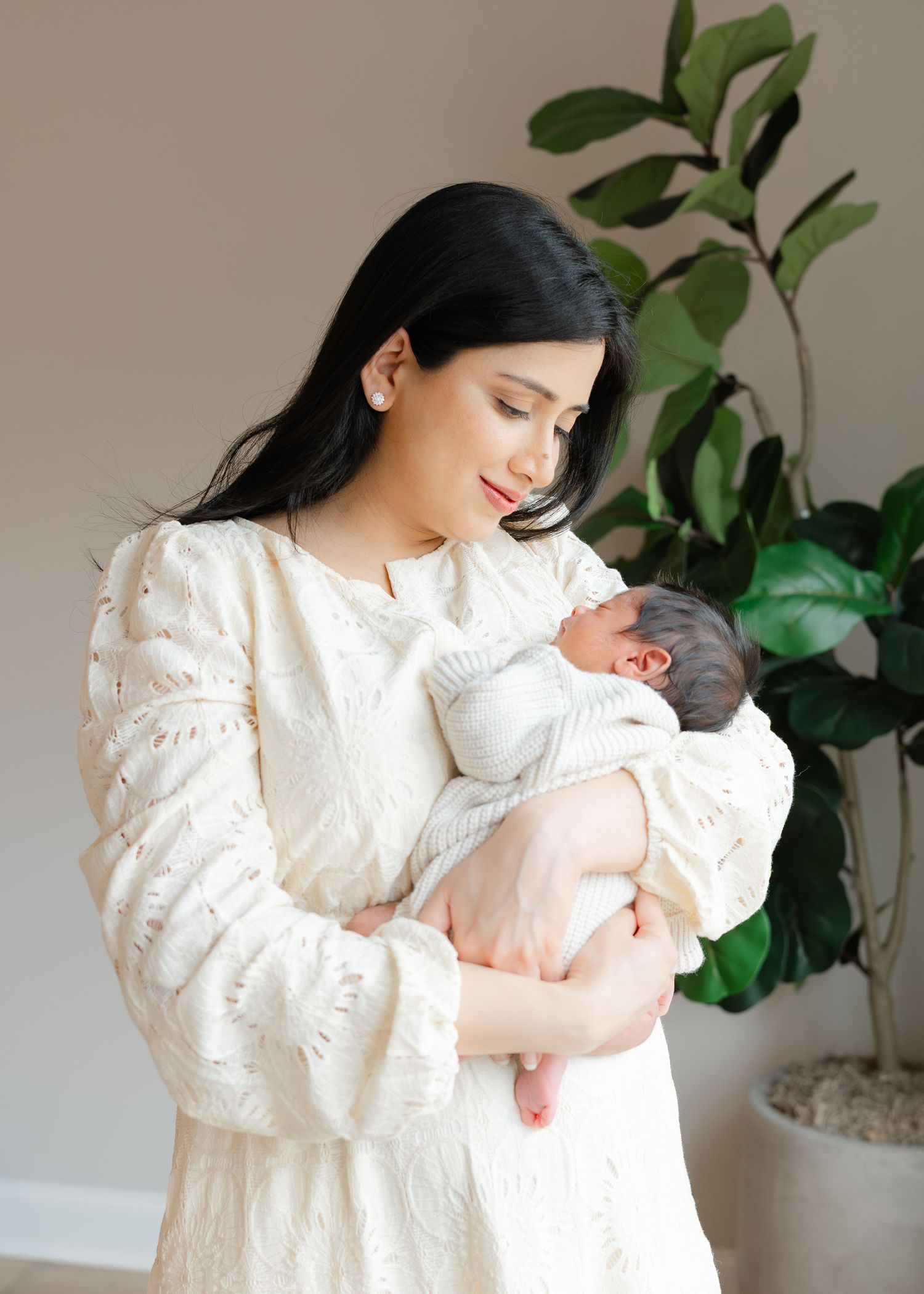 A mother in a white lace dress tenderly holds her newborn baby while standing near a fiddle leaf fig plant.