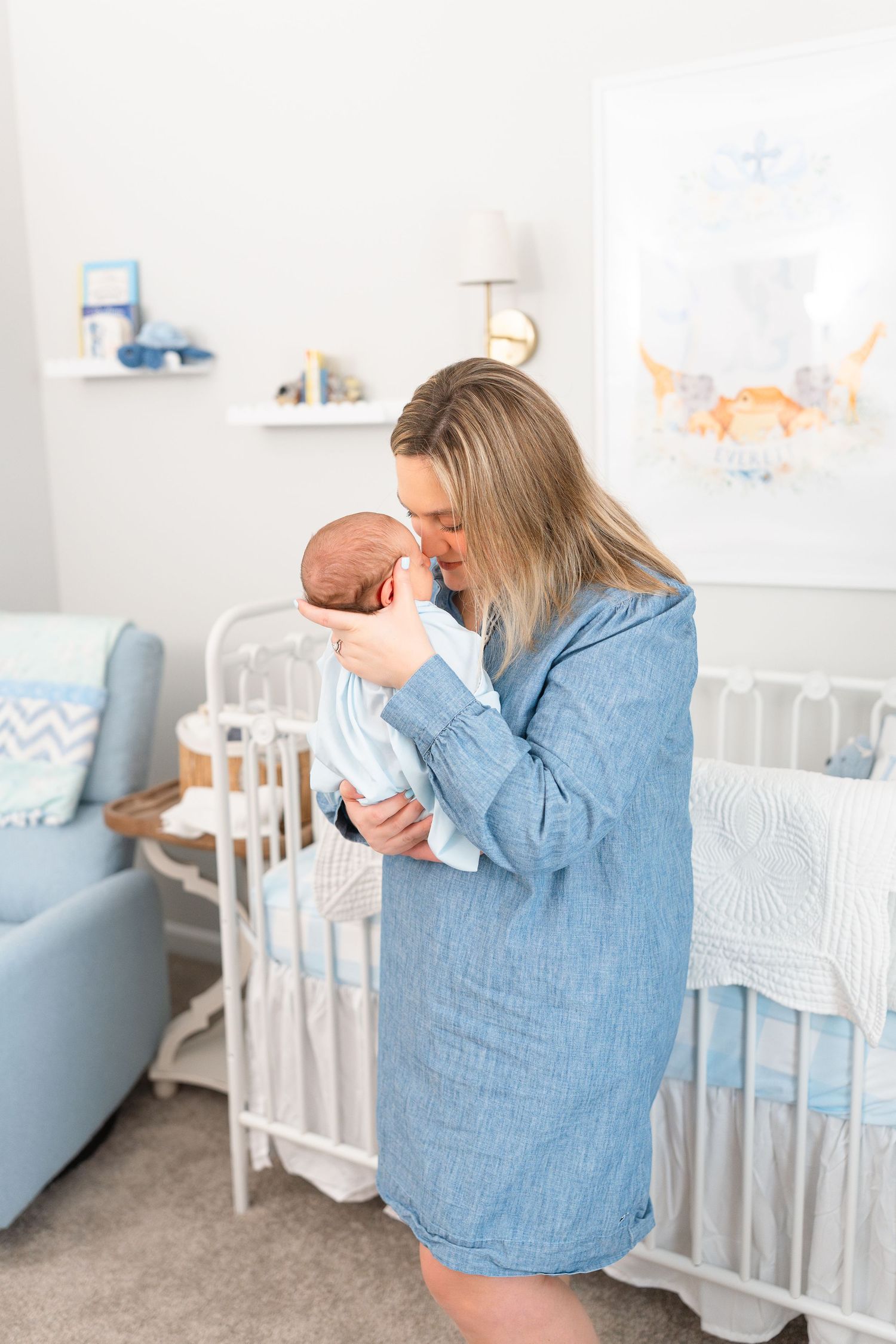 Mother in denim dress kissing newborn baby while standing in decorated blue nursery.