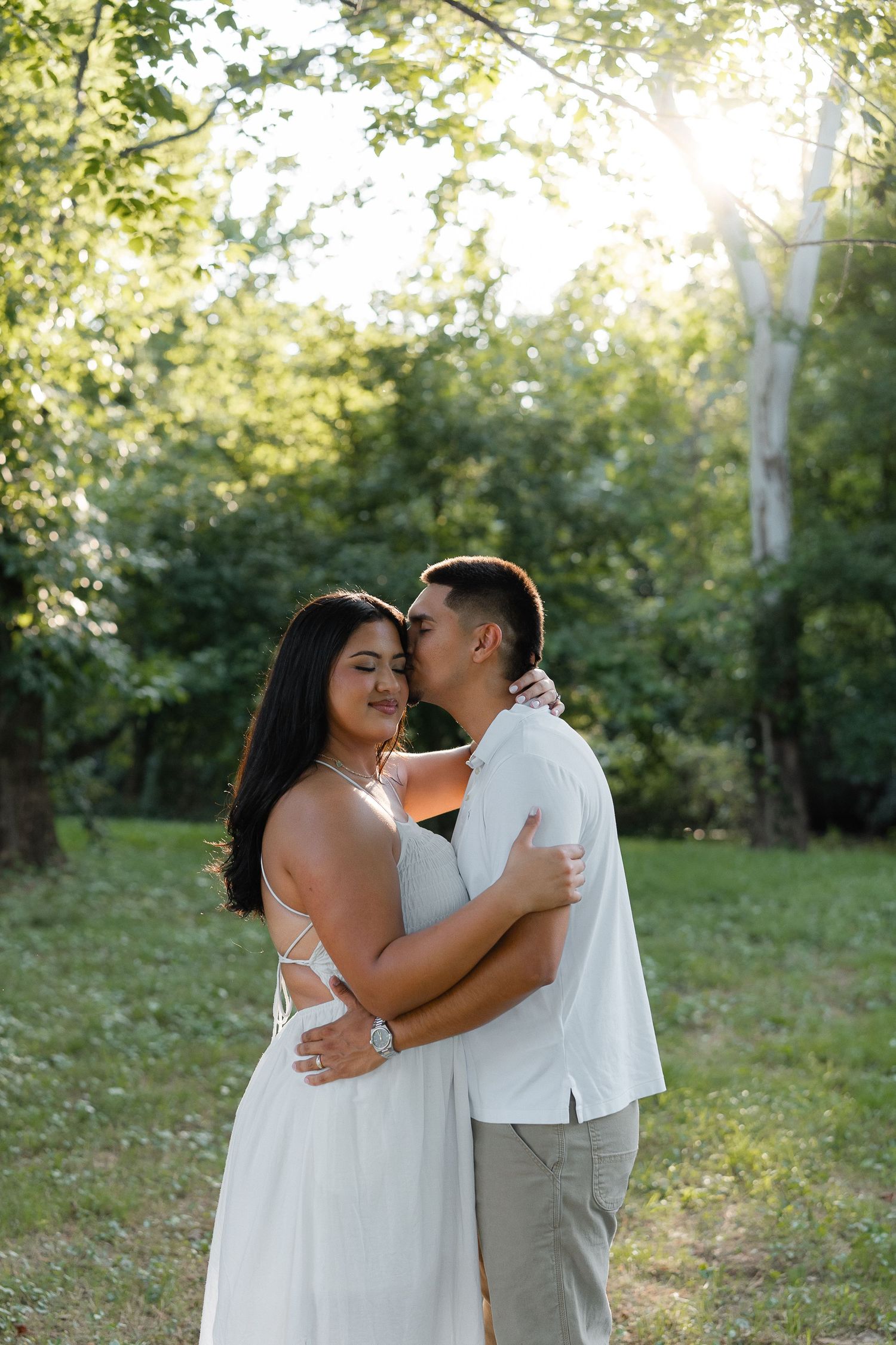 Couple embracing and kissing in a sunlit forest clearing, woman in white dress, man in white shirt and khaki pants.