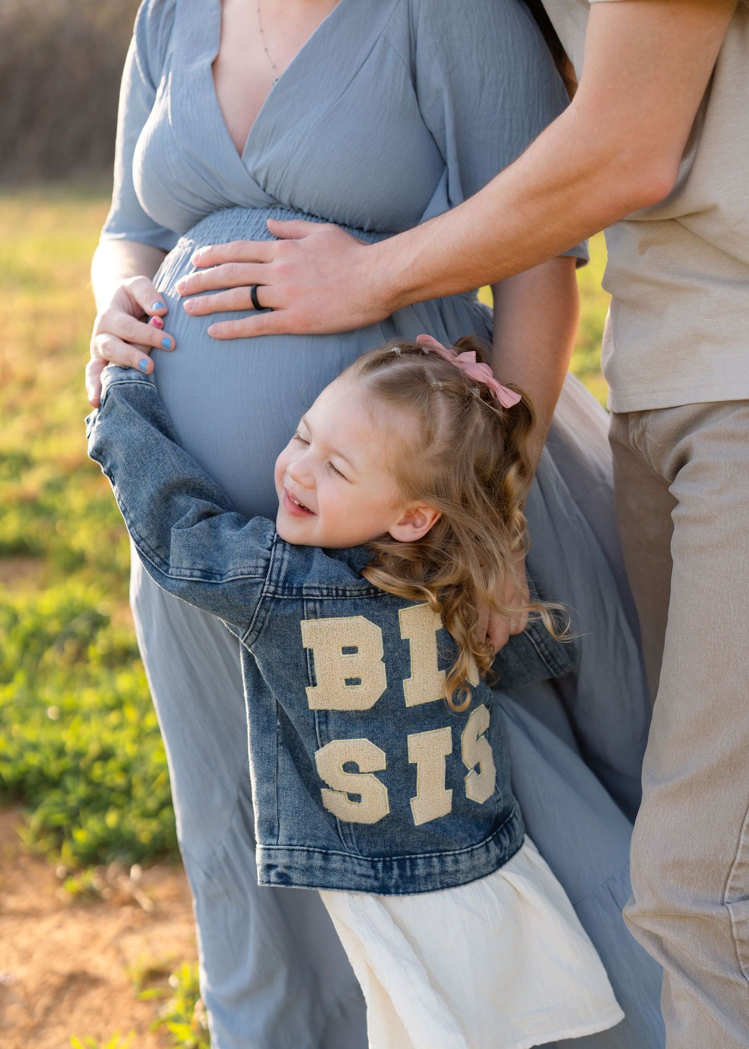 A toddler girl in a denim Big Sis jacket hugs her moms pregnant belly during an outdoor maternity photo session.