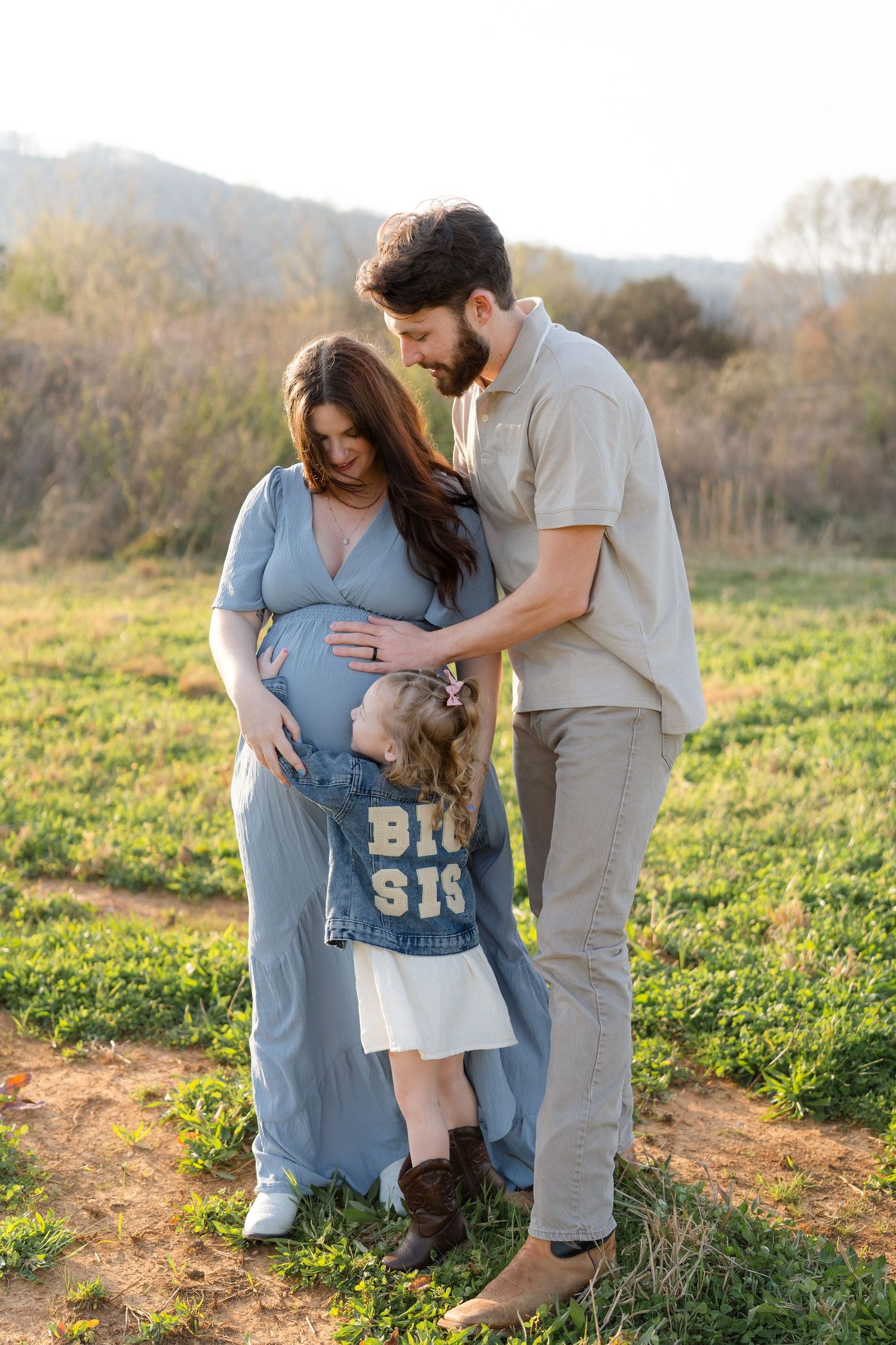 A pregnant couple in a field tenderly looks down at their young daughter wearing a Big Sis denim jacket.
