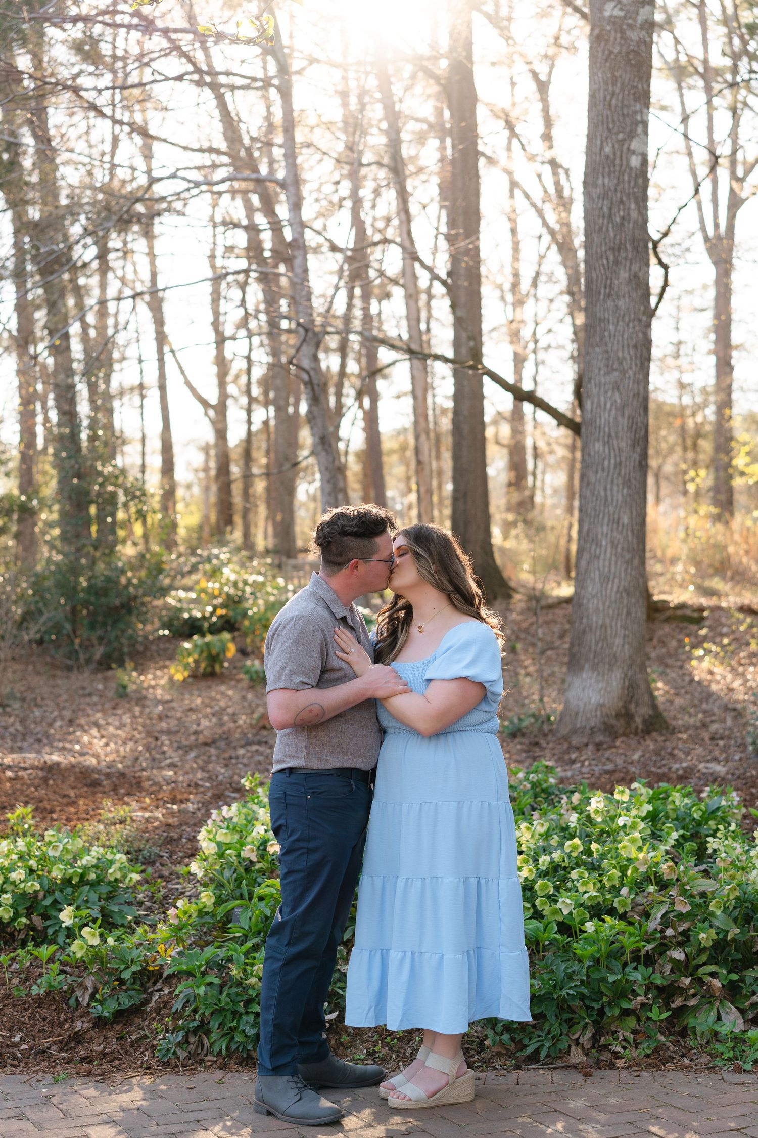 A pregnant couple shares a kiss on a sunlit garden path surrounded by tall trees and green foliage.