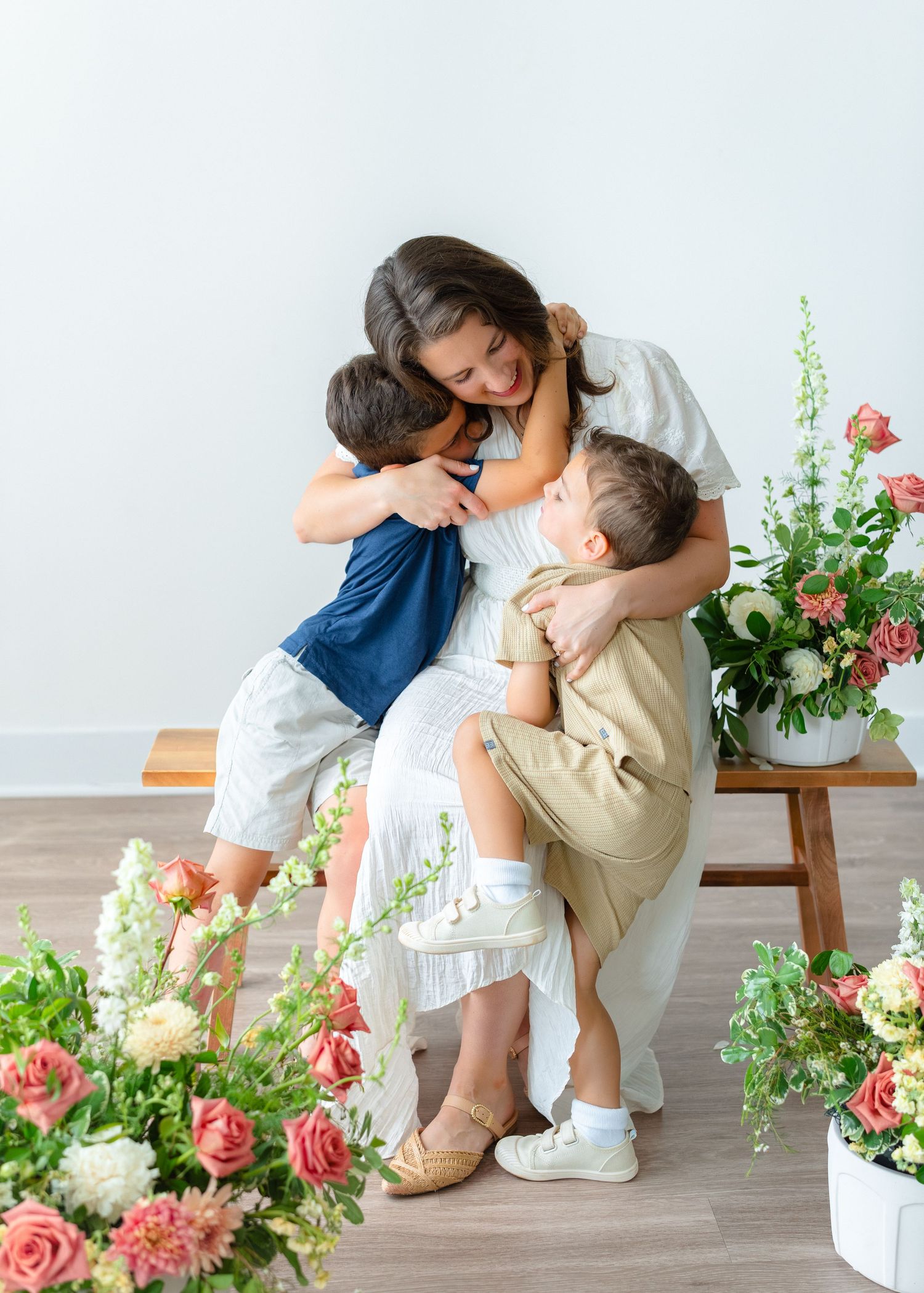 Two young boys embrace their smiling mother in a white dress surrounded by floral arrangements.