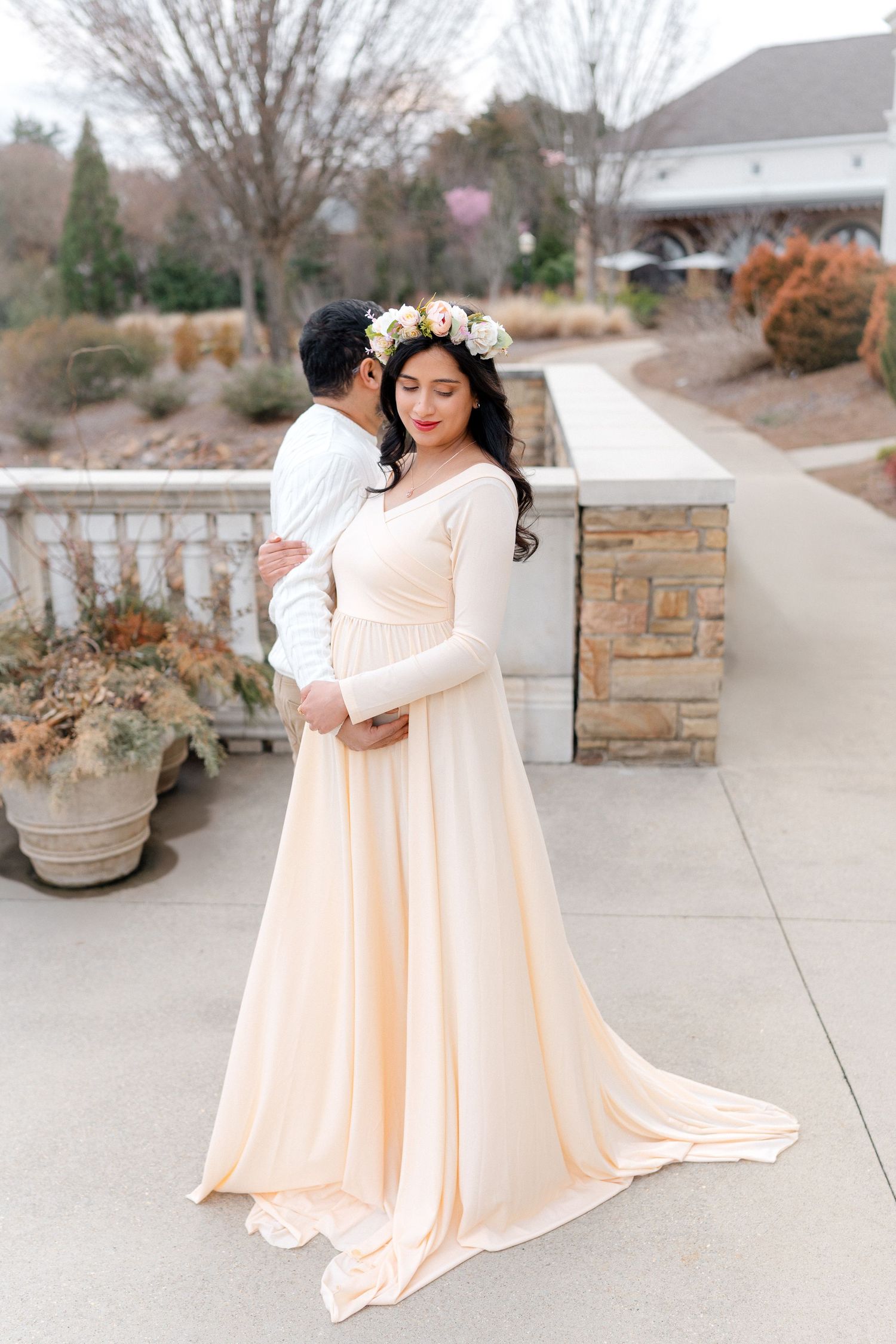 Couple embracing outdoors, woman in flowing cream maternity gown and floral crown.