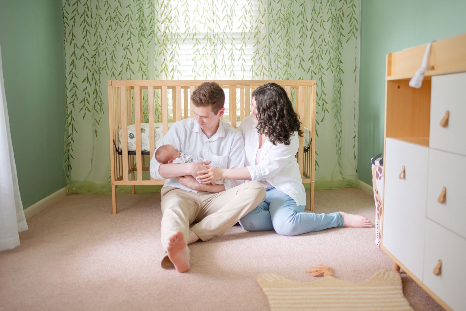 New parents sitting on nursery floor with newborn baby near wooden crib and green decor.