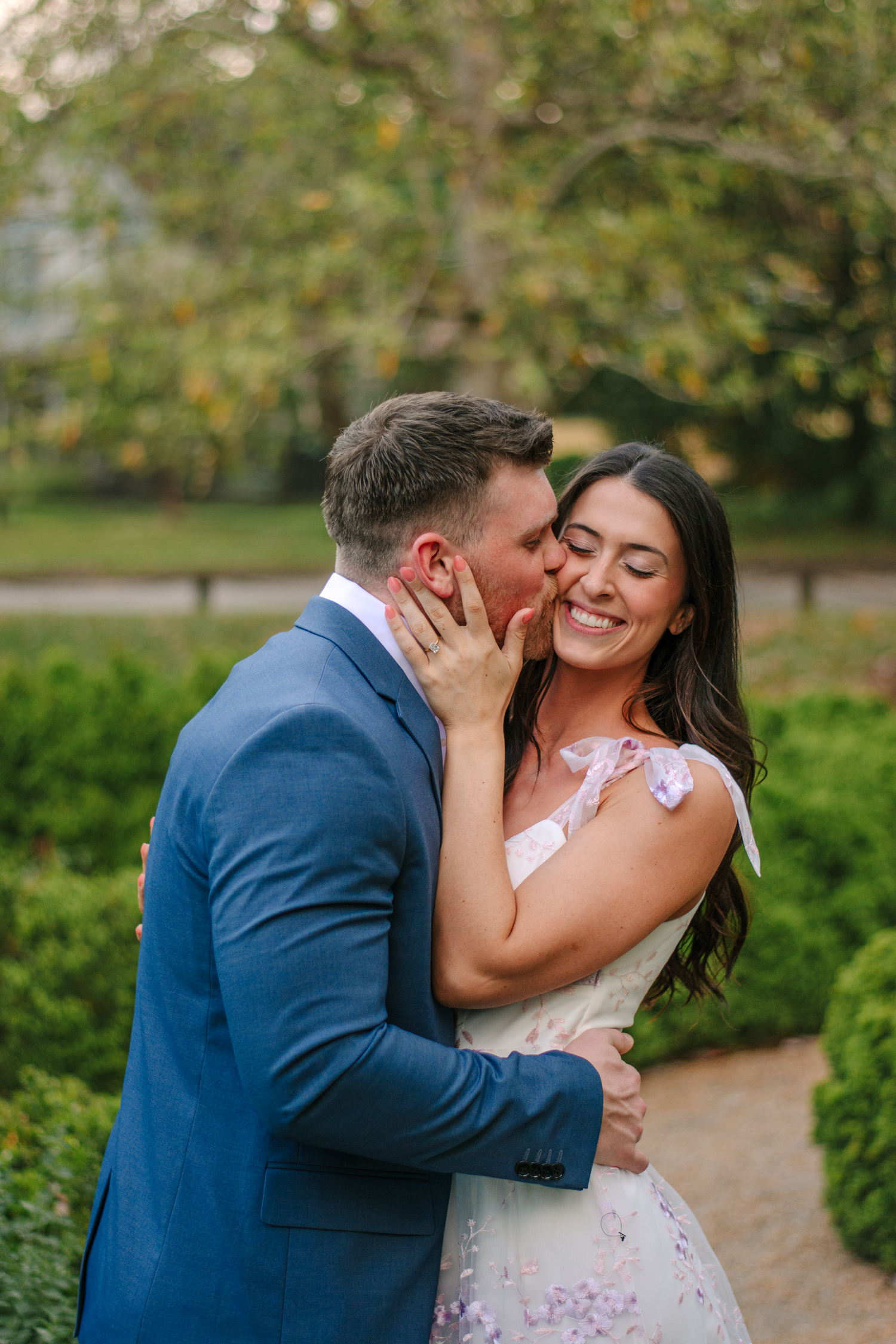 Romantic couple sharing a kiss in a garden, groom in blue suit, bride in floral white dress, smiling joyfully.