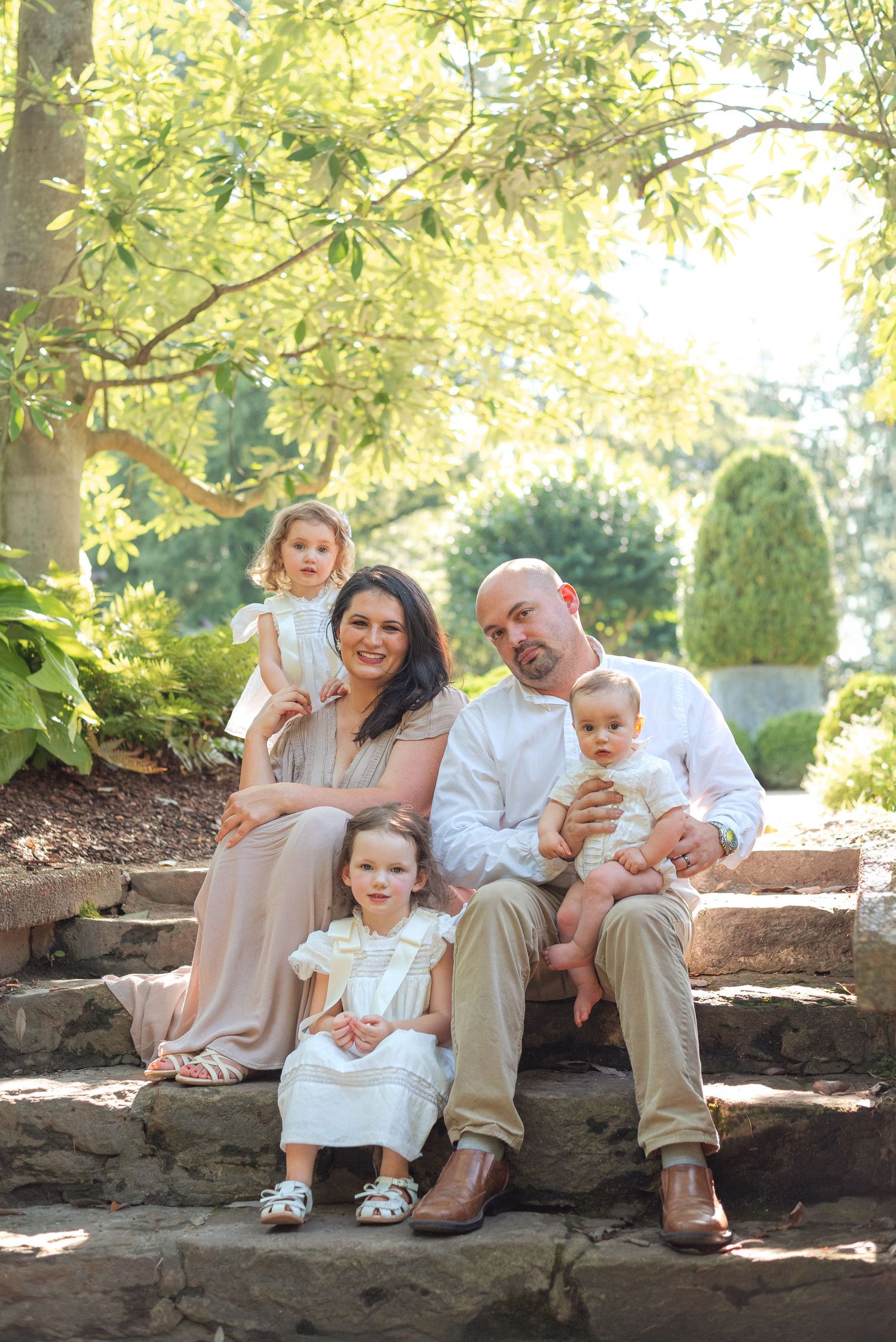 Family of five poses on stone steps surrounded by lush greenery in a sunlit garden.