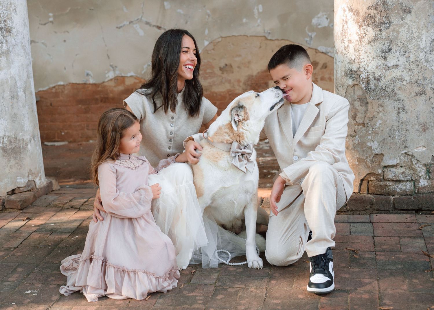 A smiling woman and two children interact with a white dog against a rustic brick wall backdrop.