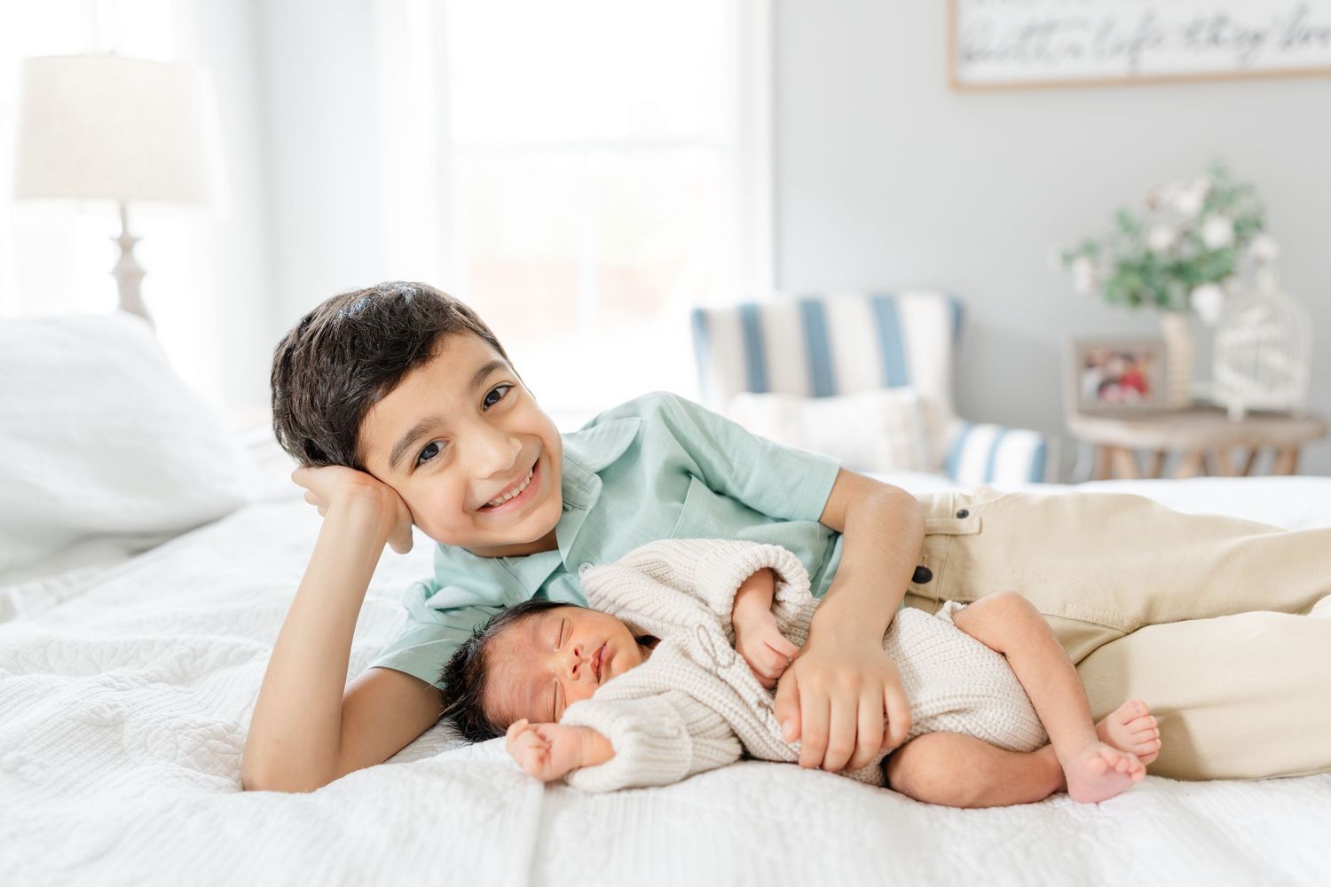 Smiling older brother lies on bed posing proudly next to sleeping newborn sibling.