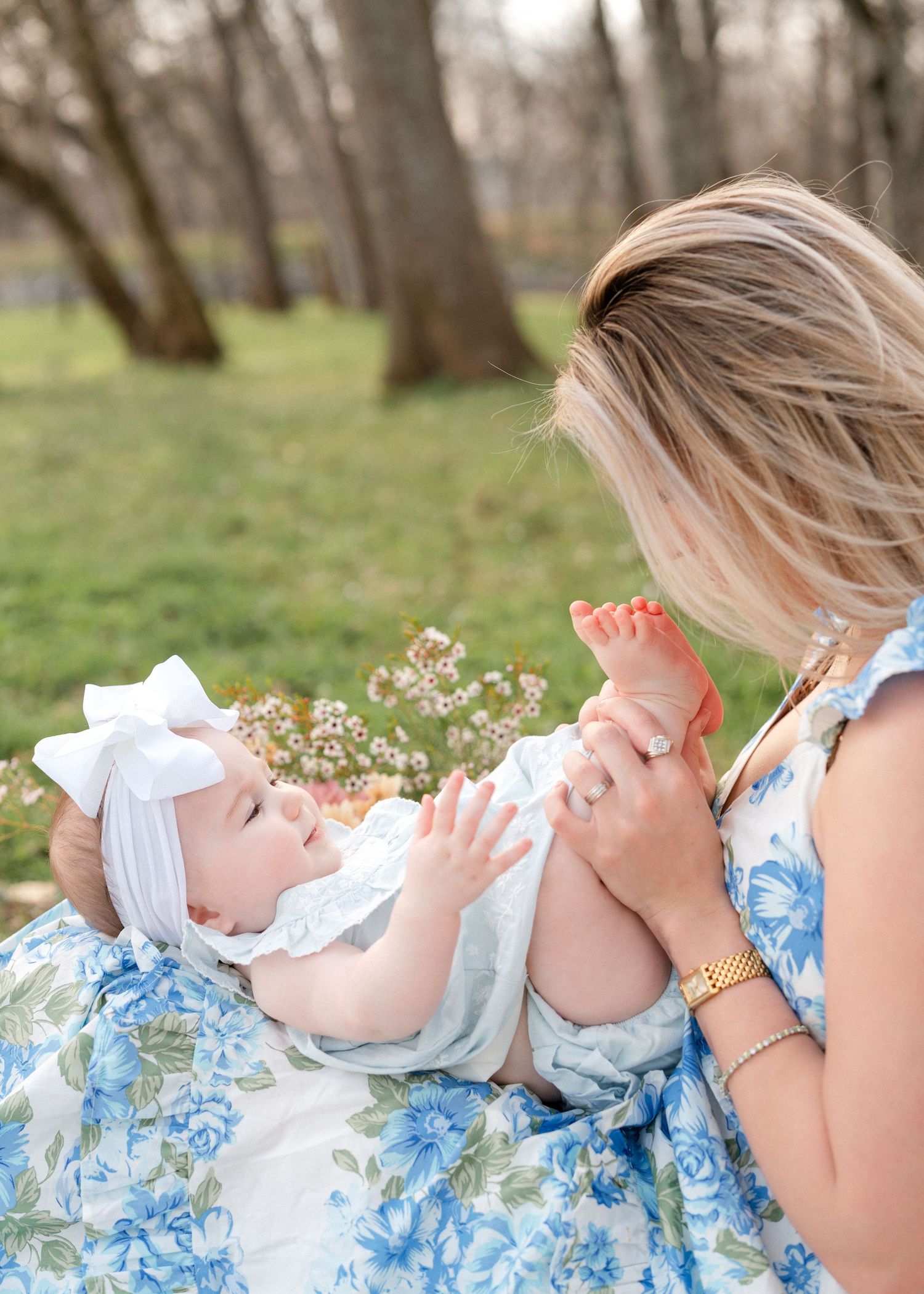 Mother in blue floral dress plays with baby girl wearing white bow headband outdoors in spring park.