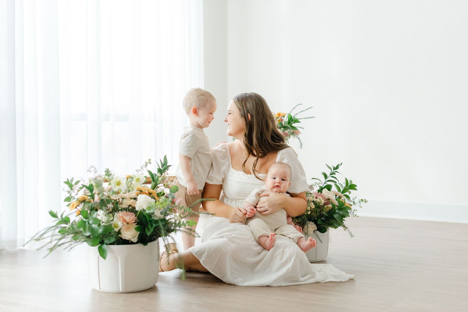 Mother in white dress sits on floor with two young children surrounded by pastel floral arrangements.