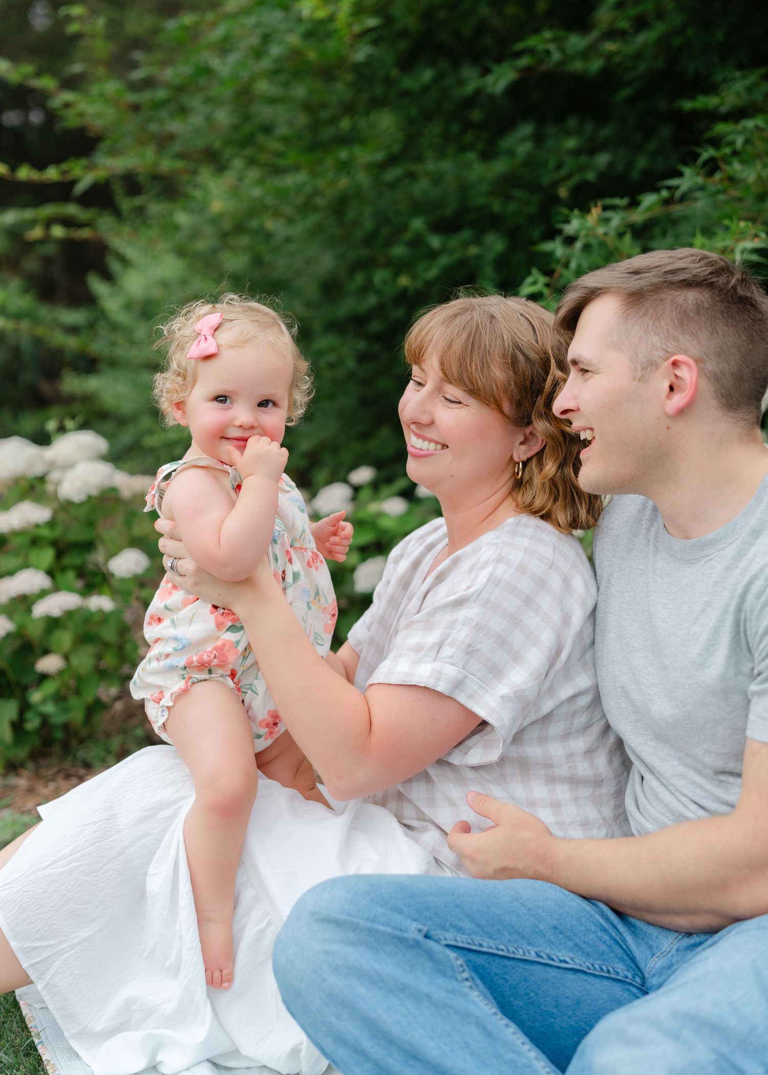 A happy family of three sits outdoors, mom holding a smiling toddler girl in a floral outfit near white hydrangeas.