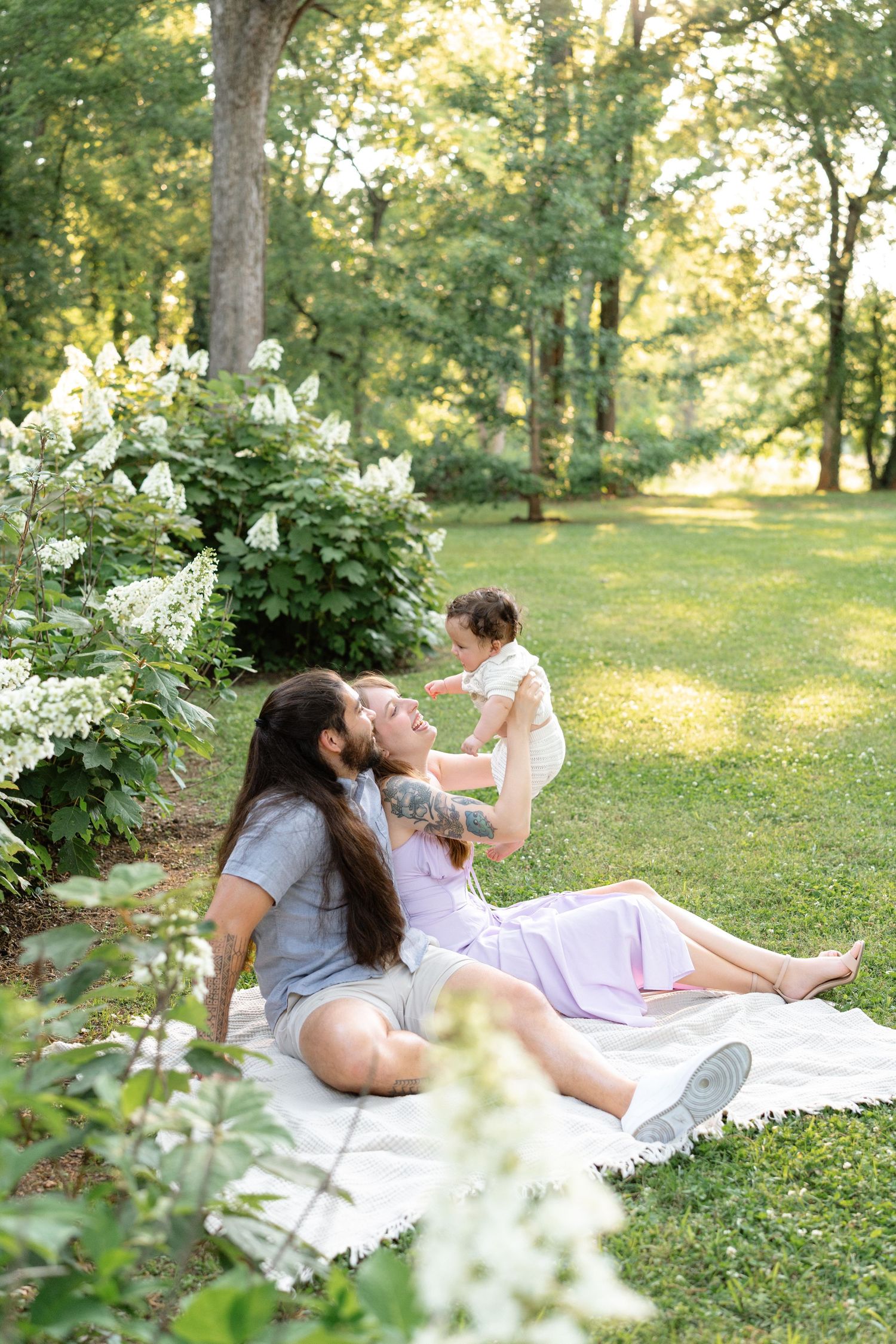 Family enjoying a sunny outdoor picnic on white blanket near blooming white hydrangeas in lush green park.