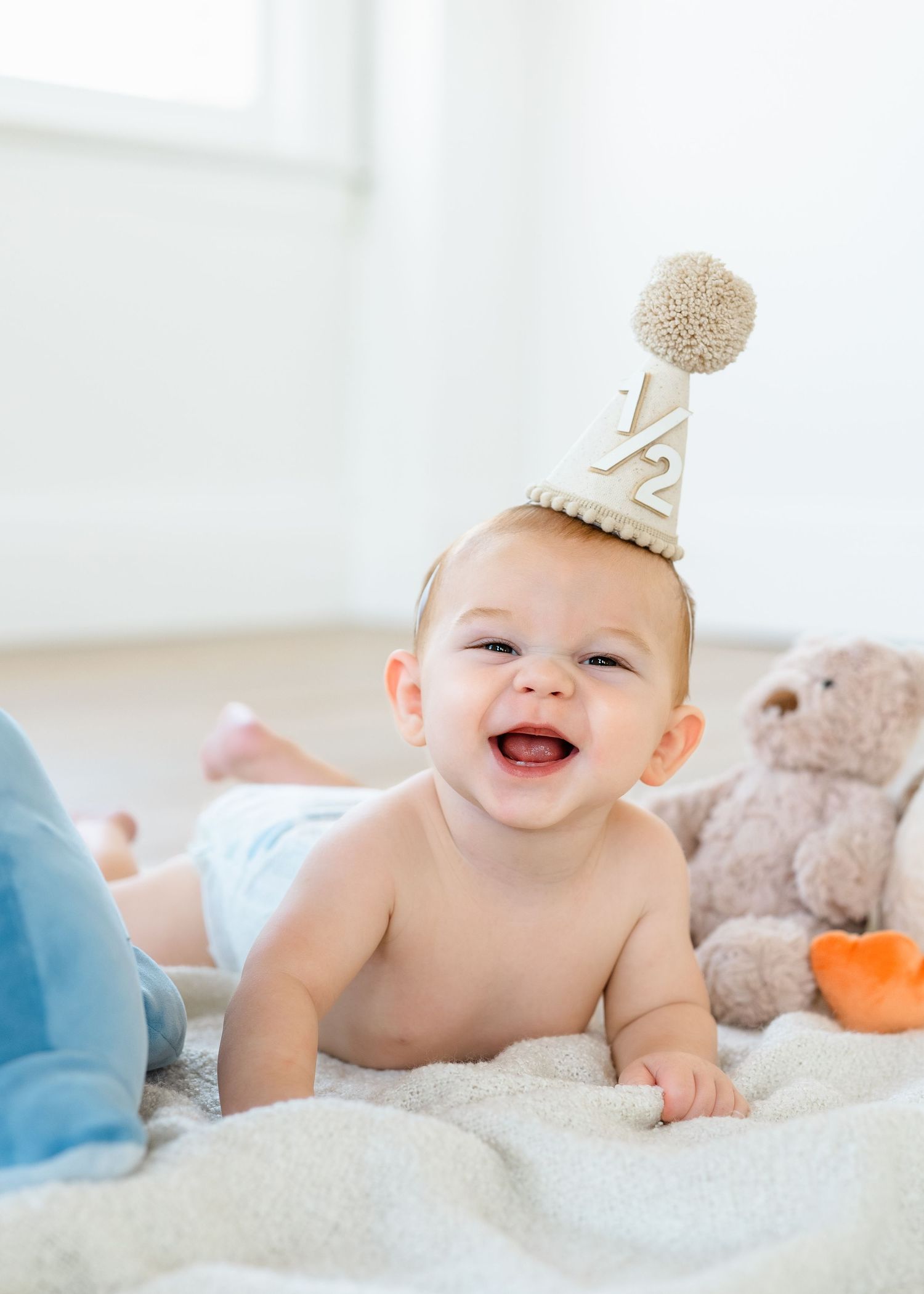 Smiling baby wearing a half birthday party hat lies on a blanket next to a teddy bear.