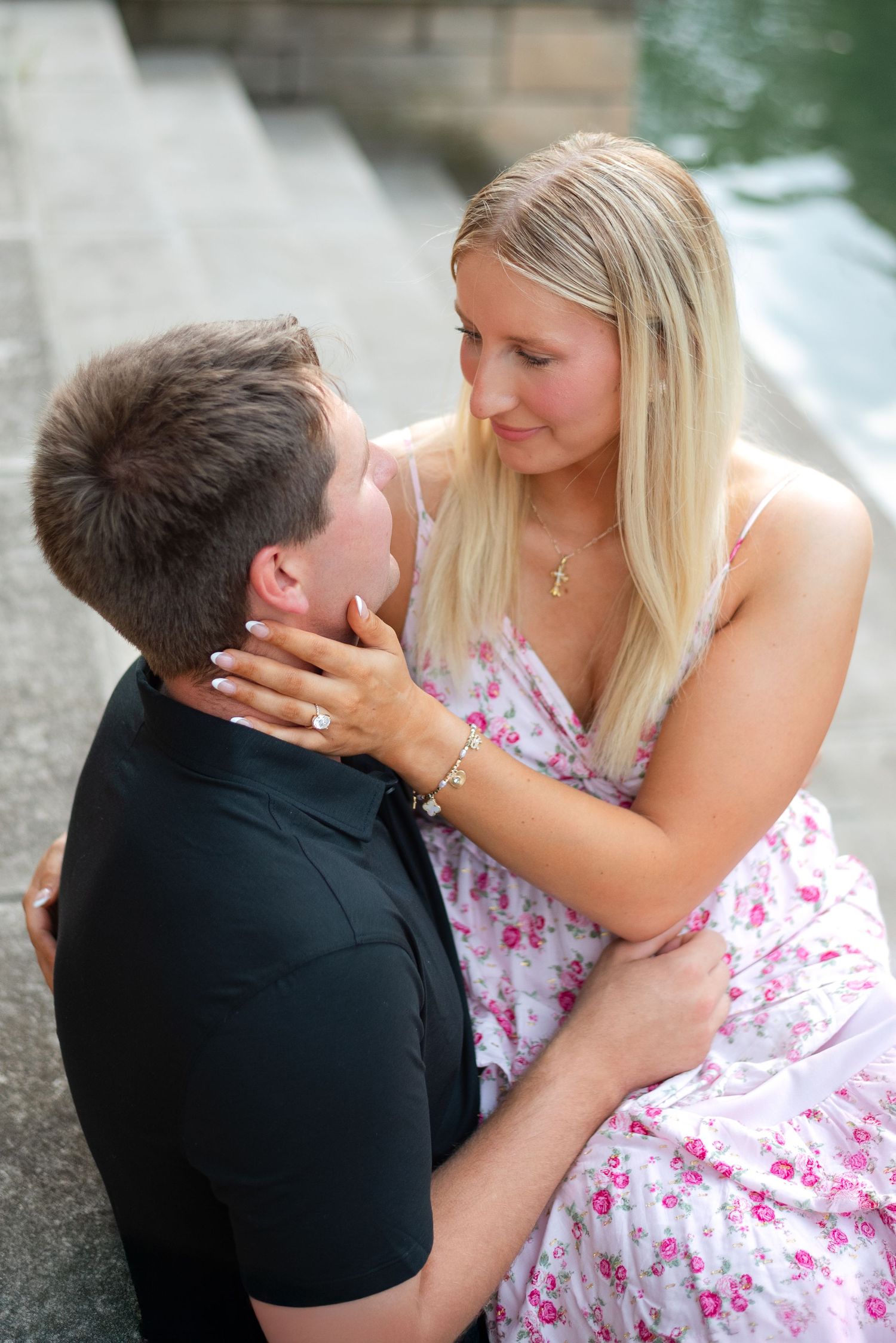 A blonde woman in a floral dress gazes at her partner during an outdoor engagement session.